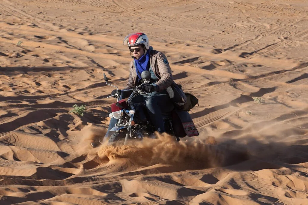 Person riding a motorcycle through a desert with sand dunes, wearing a helmet, sunglasses, and a jacket.