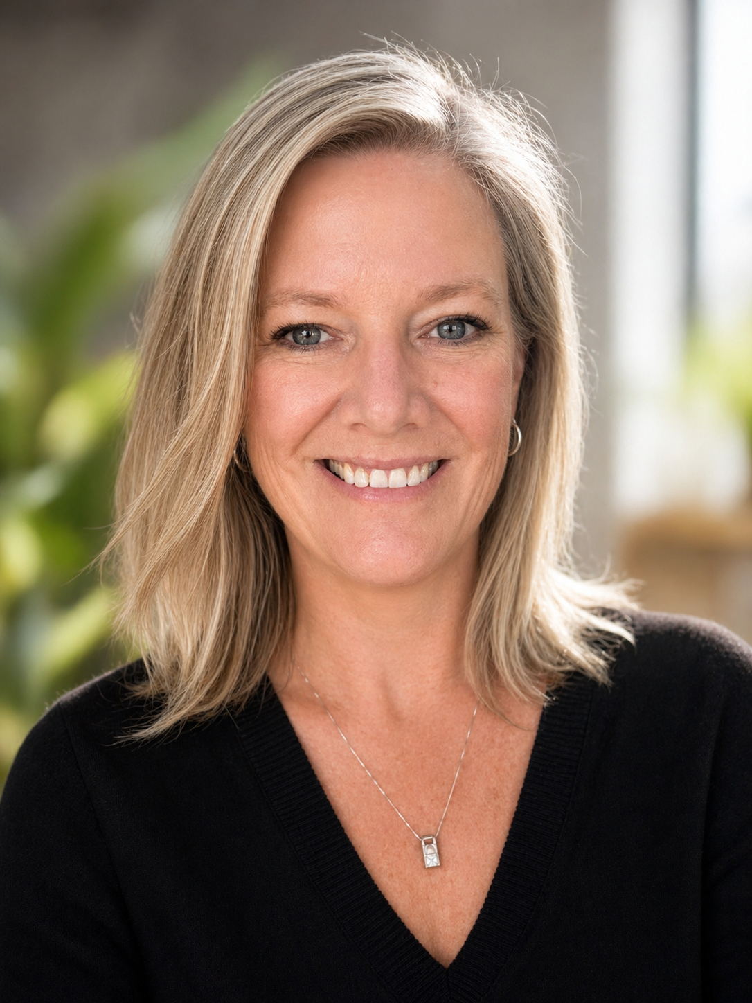 A woman with shoulder-length blonde hair, blue eyes, and a smile, wearing a black top and silver necklace, standing indoors with a blurred background of green plants and sunlight.