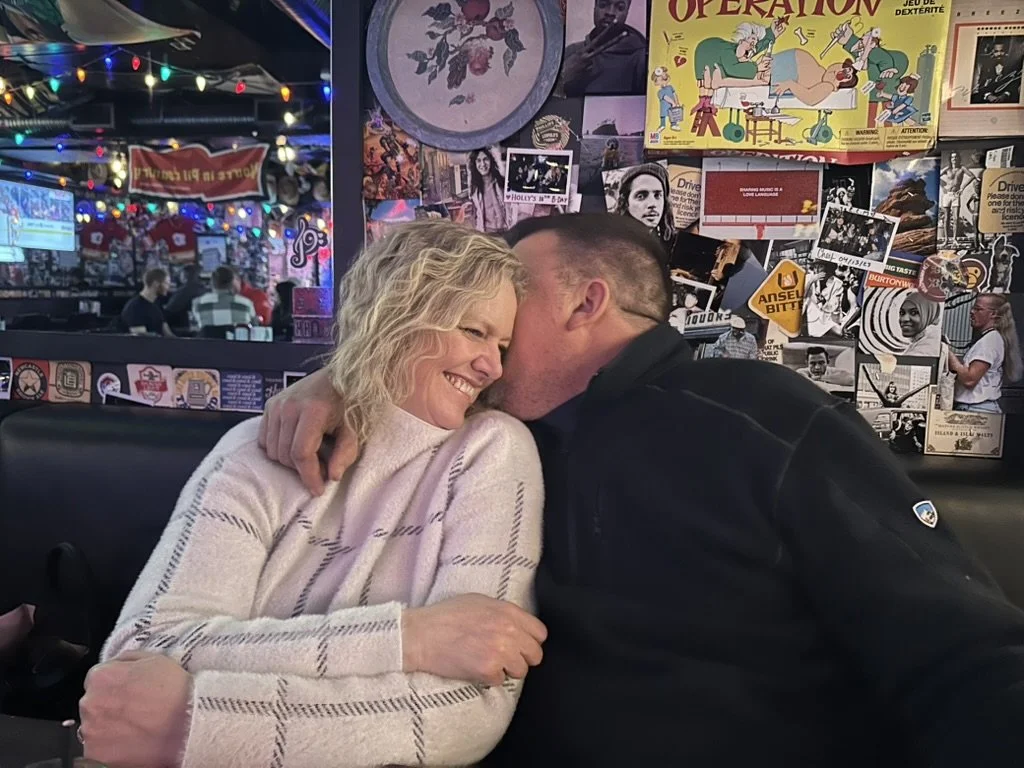 A couple sitting close together in a restaurant or bar, with the woman smiling and the man leaning in for a kiss. The background features vibrant wall art, posters, and photos, with colorful string lights overhead.