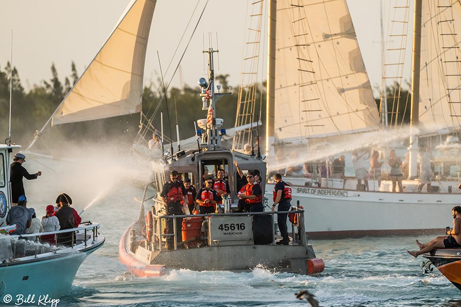 A busy water scene with sailboats and smaller motorboats, including life jackets on some people and water spray, possibly during a regatta or sailing event.