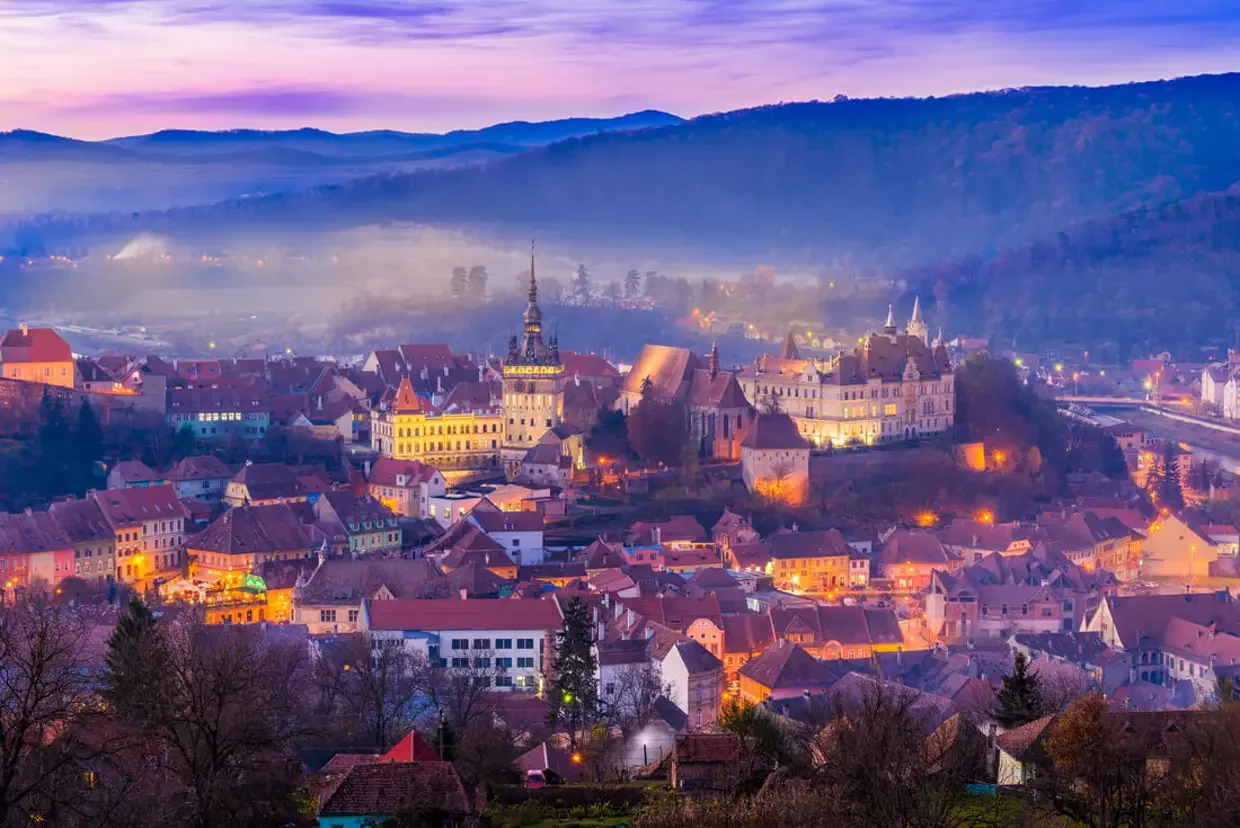 A panoramic view of a European town at dusk with illuminated buildings, a tall church spire, rolling hills in the background, and a misty atmosphere.