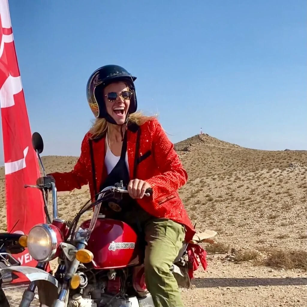 Woman wearing a helmet and sunglasses, laughing, riding a red motorcycle in a desert landscape with a hill in the background.