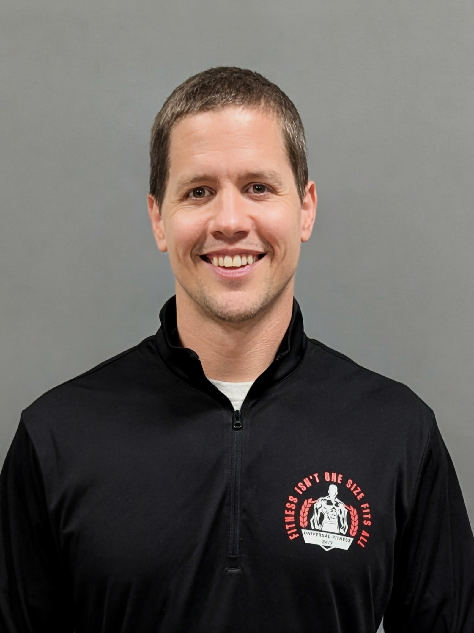 A smiling man with short brown hair and wearing a black zip-up jacket with a logo on the front, standing against a gray background.