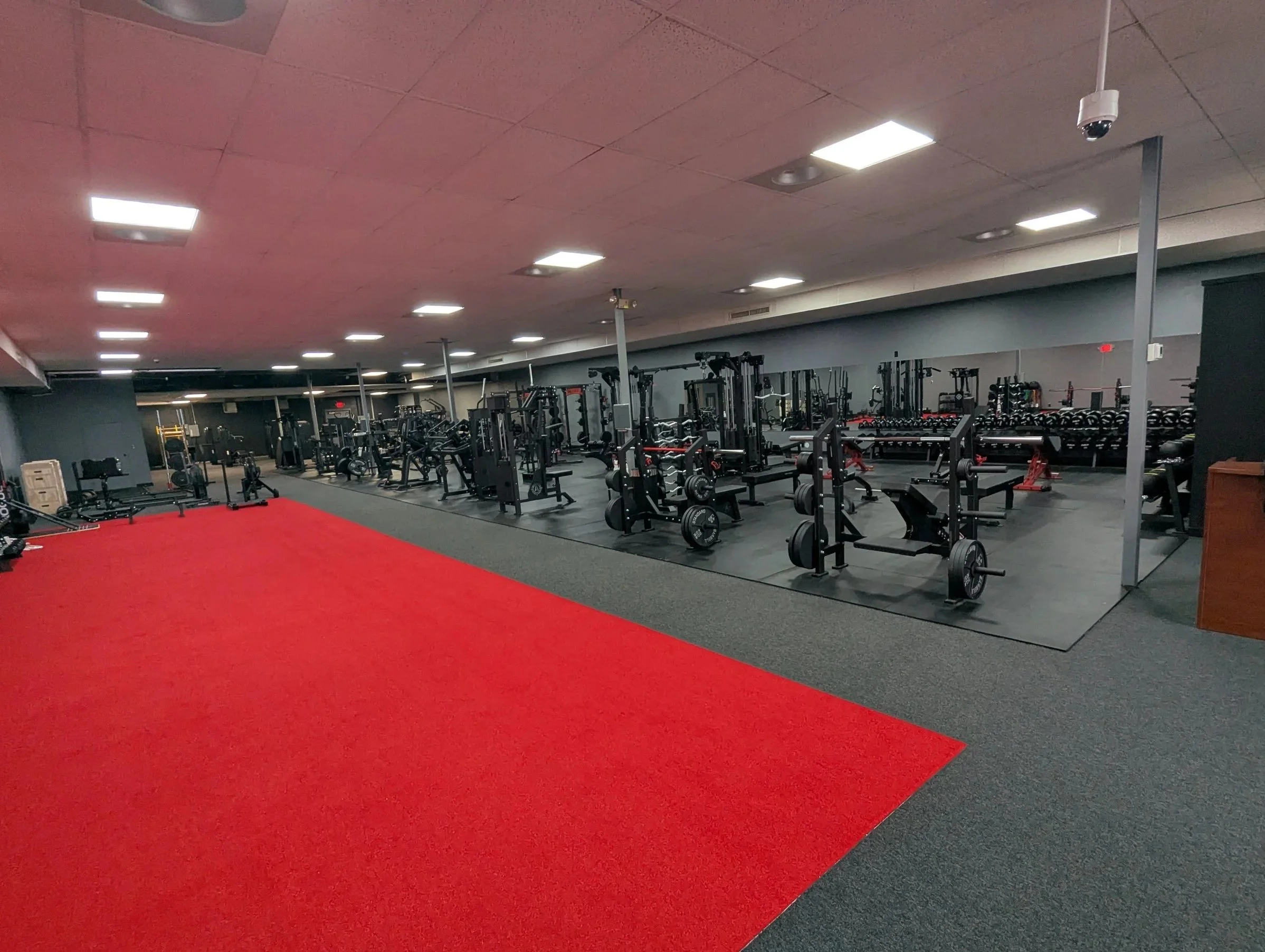 A gym with a red mat on the floor and various weightlifting equipment, including barbells and racks, in the background under bright ceiling lights.