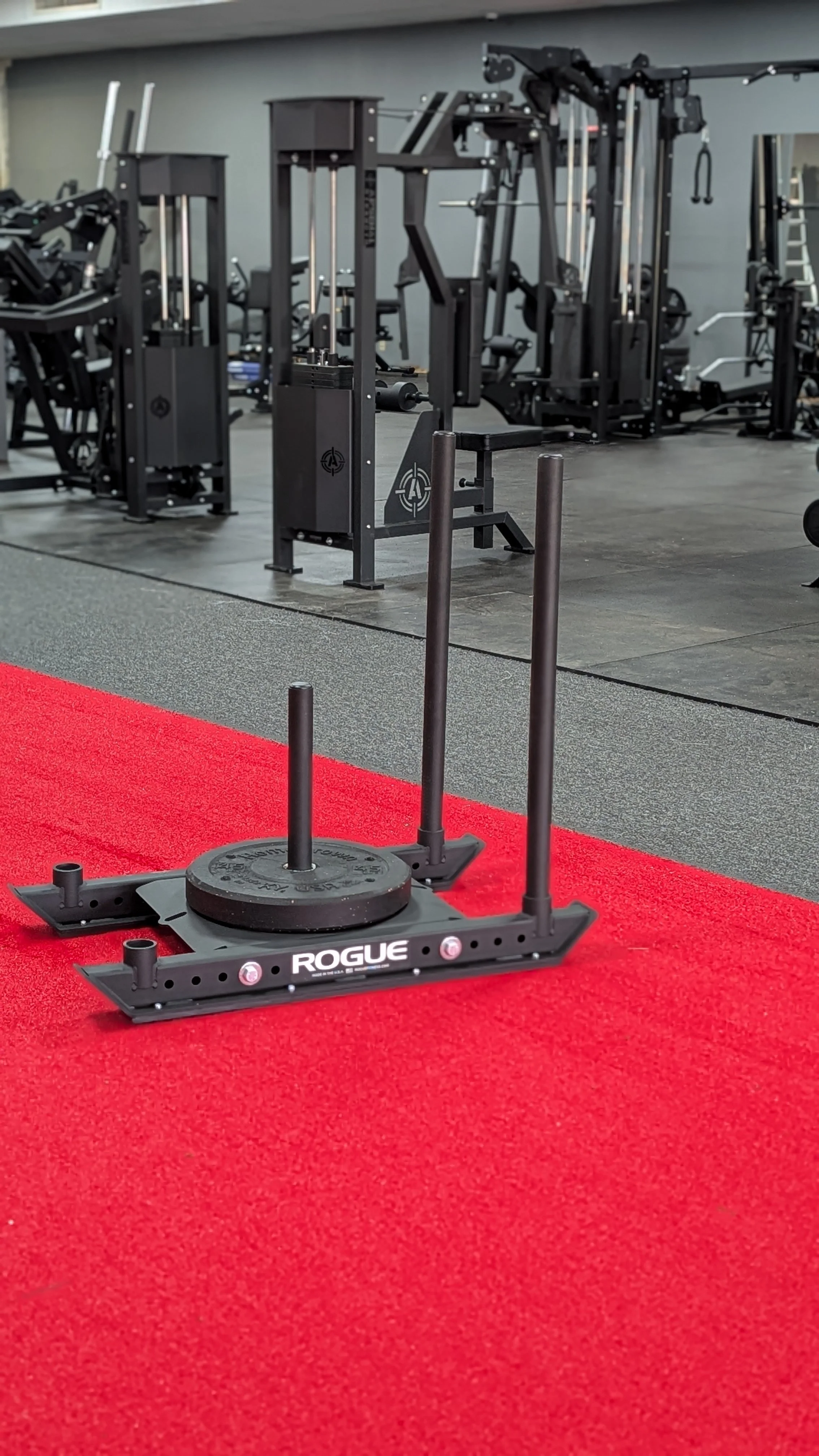 Indoor gym with black exercise equipment, a red workout mat, and a bumper plate with a sled and weight plates in the foreground.