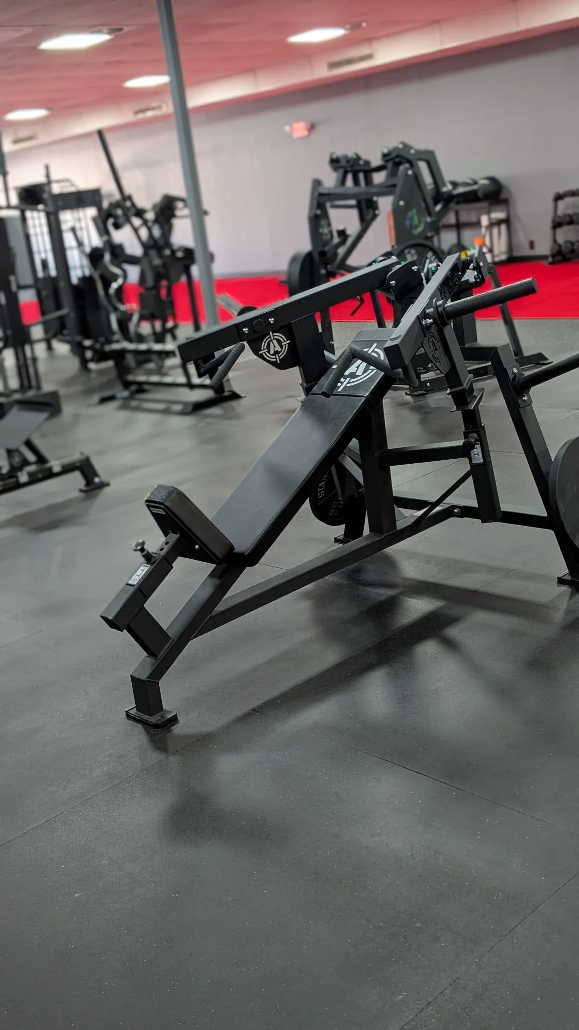 Empty gym with various workout equipment, including benches, weight machines, and dumbbells, on black floor with red carpeting in the background.