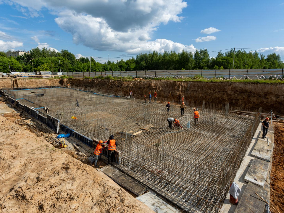 Construction workers install rebar in a large excavation site under a partly cloudy sky.