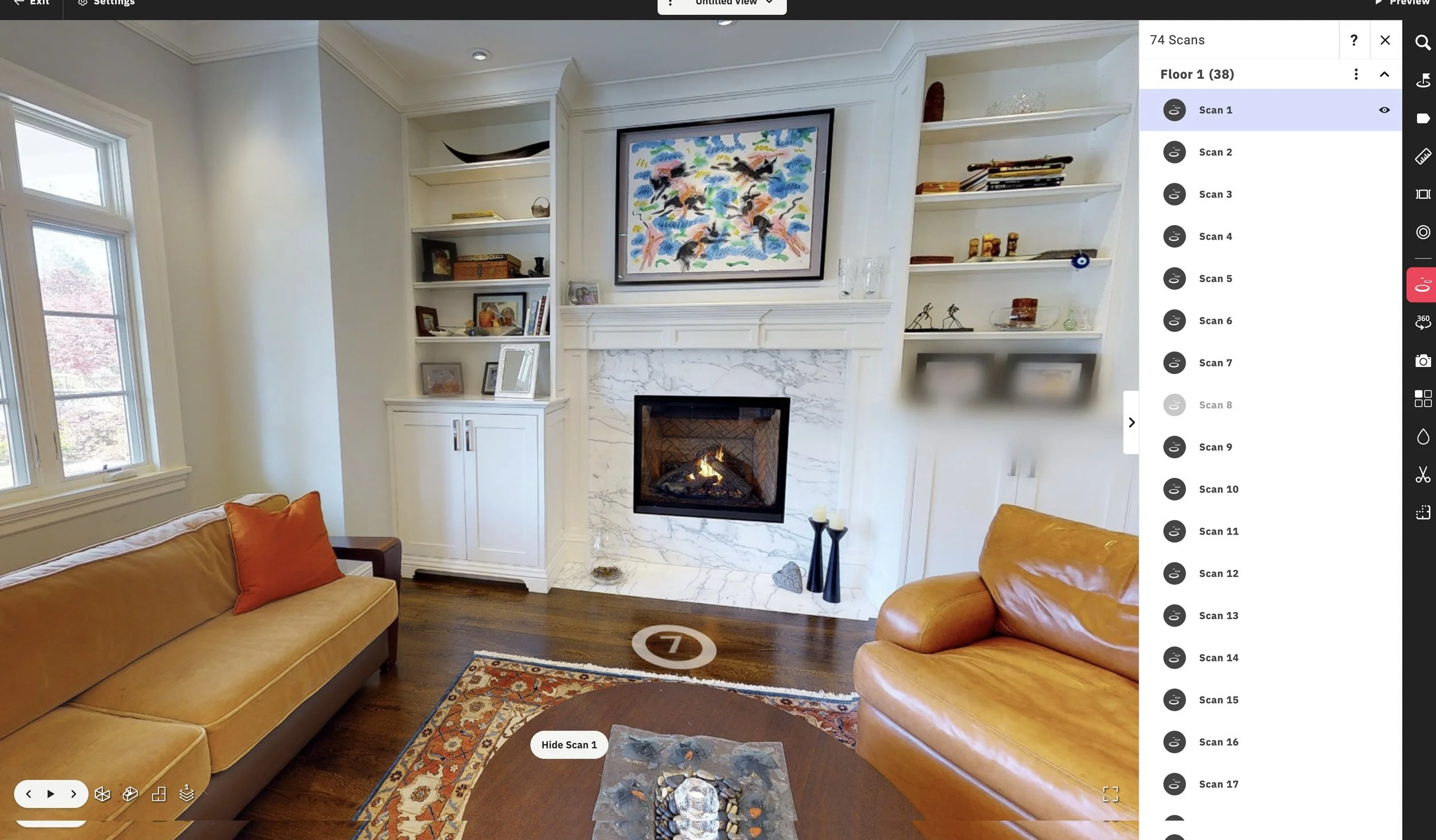 Living room with a fireplace and built-in bookshelves, decorated with books, sculptures, and framed photos. The room has a window on the left side, a tan sofa with orange pillows, and a small table with decorative items. There is colorful artwork above the fireplace and a patterned rug on the wooden floor.