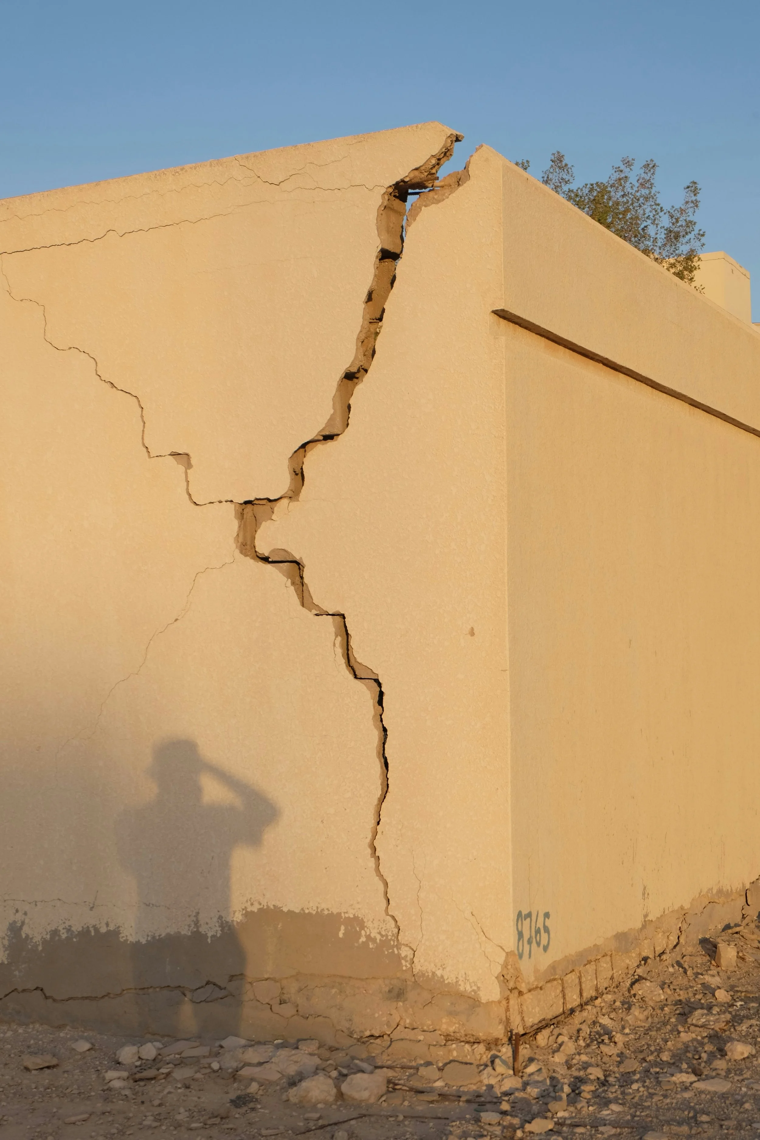 A beige concrete wall with a large crack running from top to bottom, with debris and rocks on the ground and part of a shadow in the lower left.