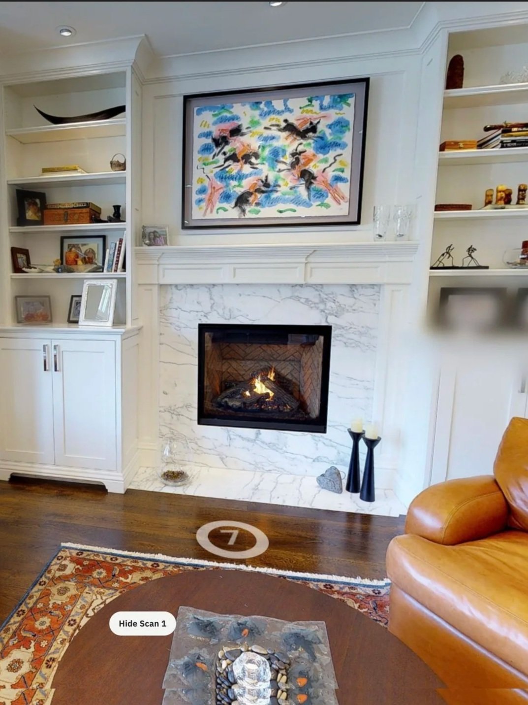 Living room with a fireplace and built-in bookshelves, decorated with books, sculptures, and framed photos. The room has a window on the left side, a tan sofa with orange pillows, and a small table with decorative items. There is colorful artwork above the fireplace and a patterned rug on the wooden floor.