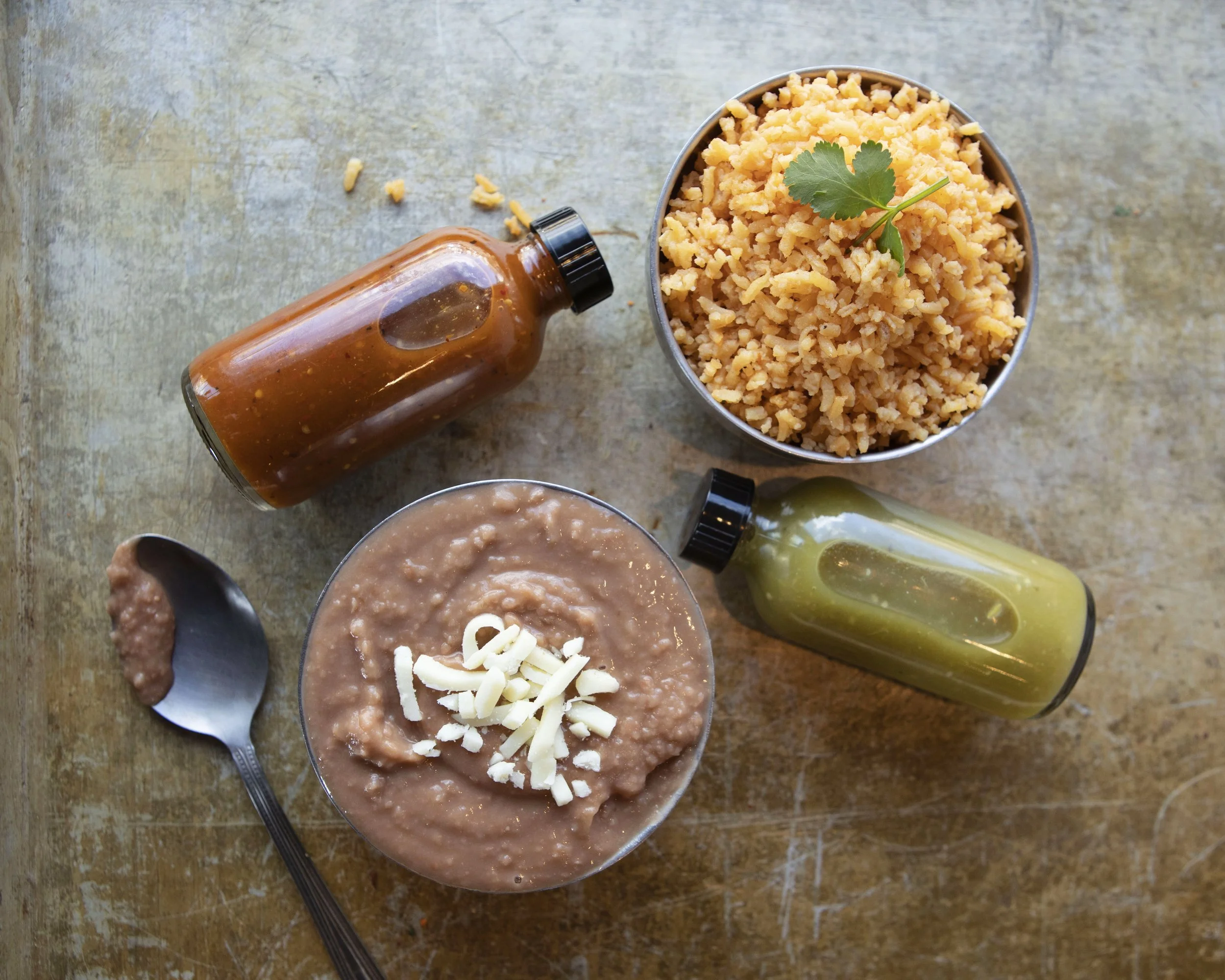 A bowl of refried beans topped with shredded cheese, a cilantro leaf, and three bottles of hot sauce on a wooden surface.