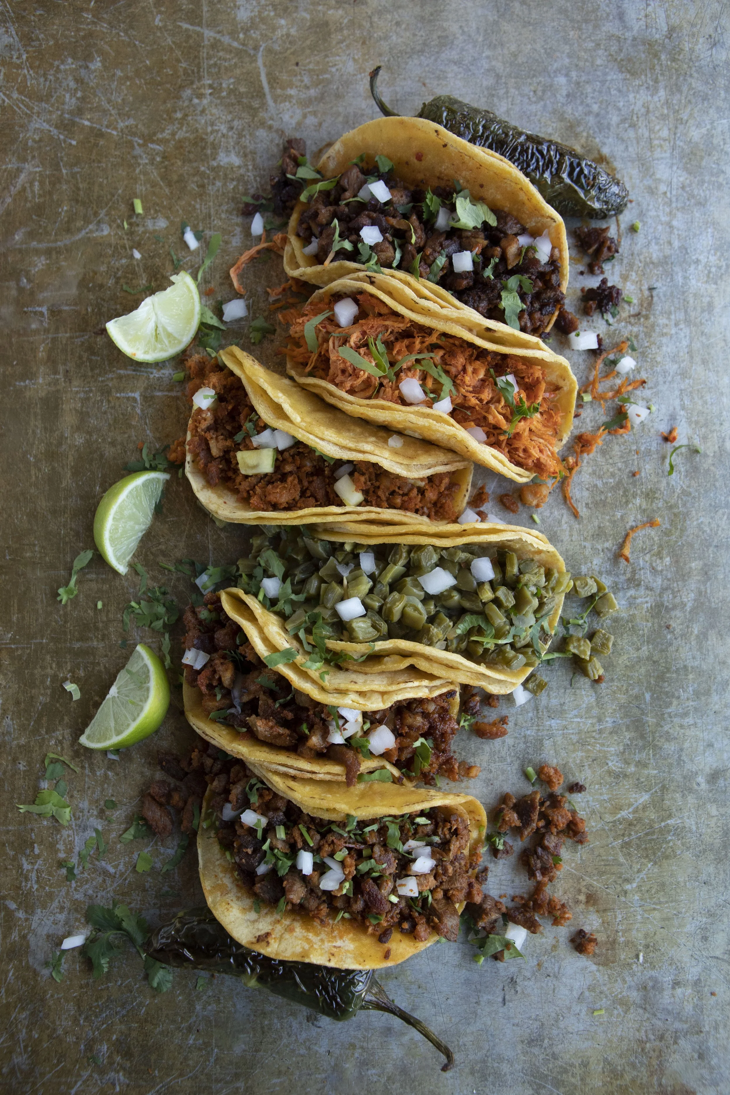 A row of five tacos filled with different meats, garnished with chopped onions and cilantro, with lime wedges and grilled peppers nearby, on a rustic metal surface.
