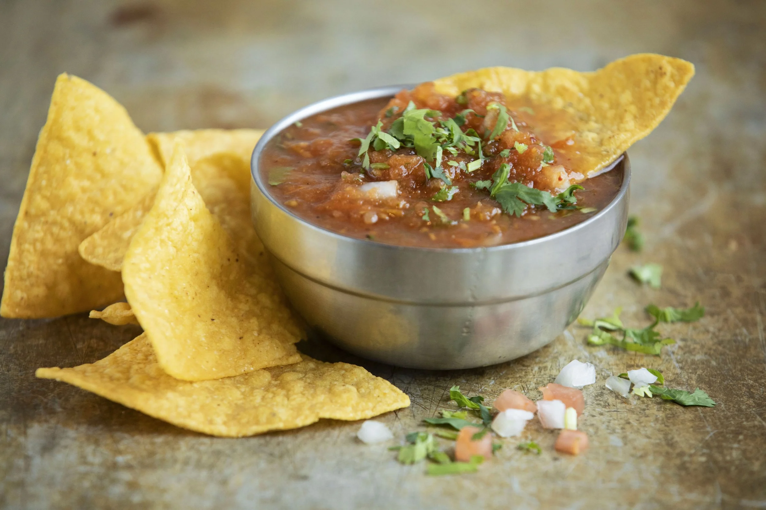 Close-up of a bowl of red chili with ground meat, topped with chopped cilantro, accompanied by yellow tortilla chips on a rustic surface with scattered chopped vegetables and herbs.