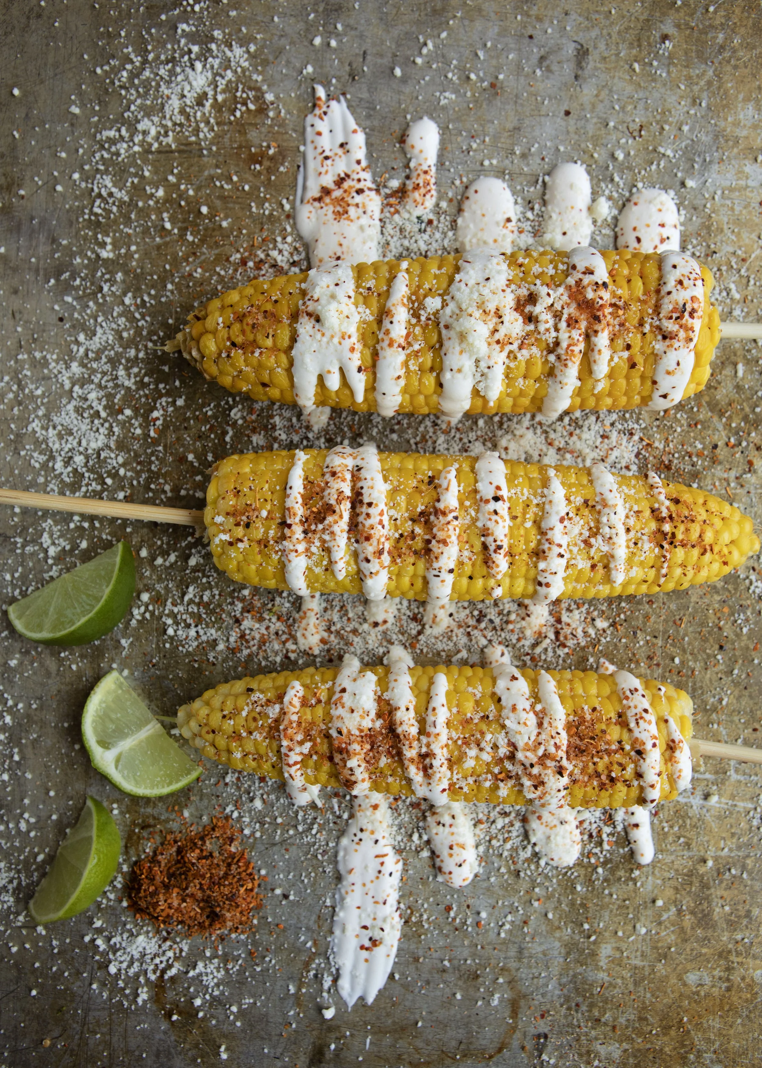 Three grilled corn on the cob skewers topped with white sauce, sprinkled with red chili powder, paprika, and black pepper, on a rustic wooden surface garnished with lime wedges and sprinkled with additional spices and salt.