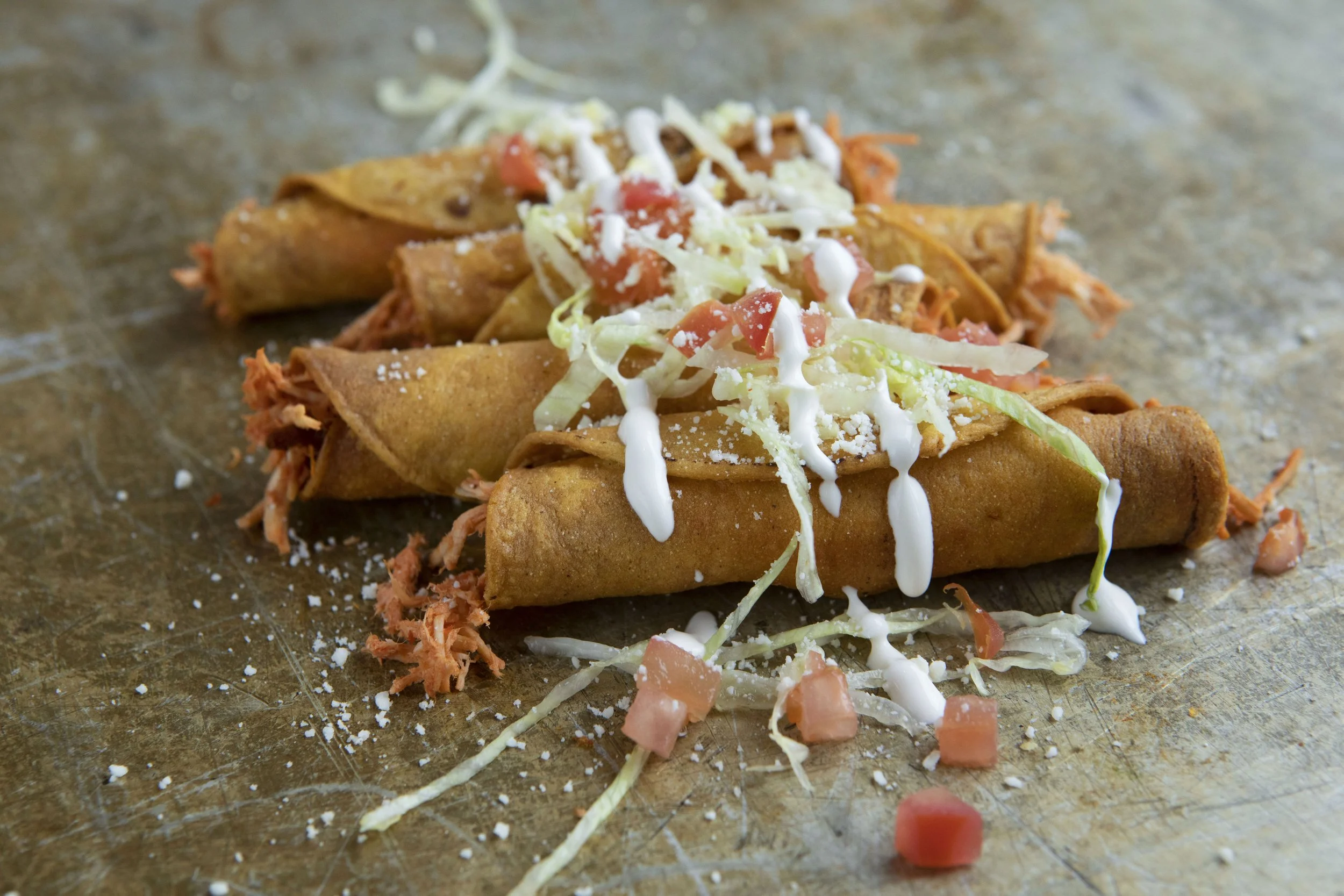 Three taquitos topped with shredded lettuce, diced tomatoes, shredded cheese, and drizzled with sour cream 