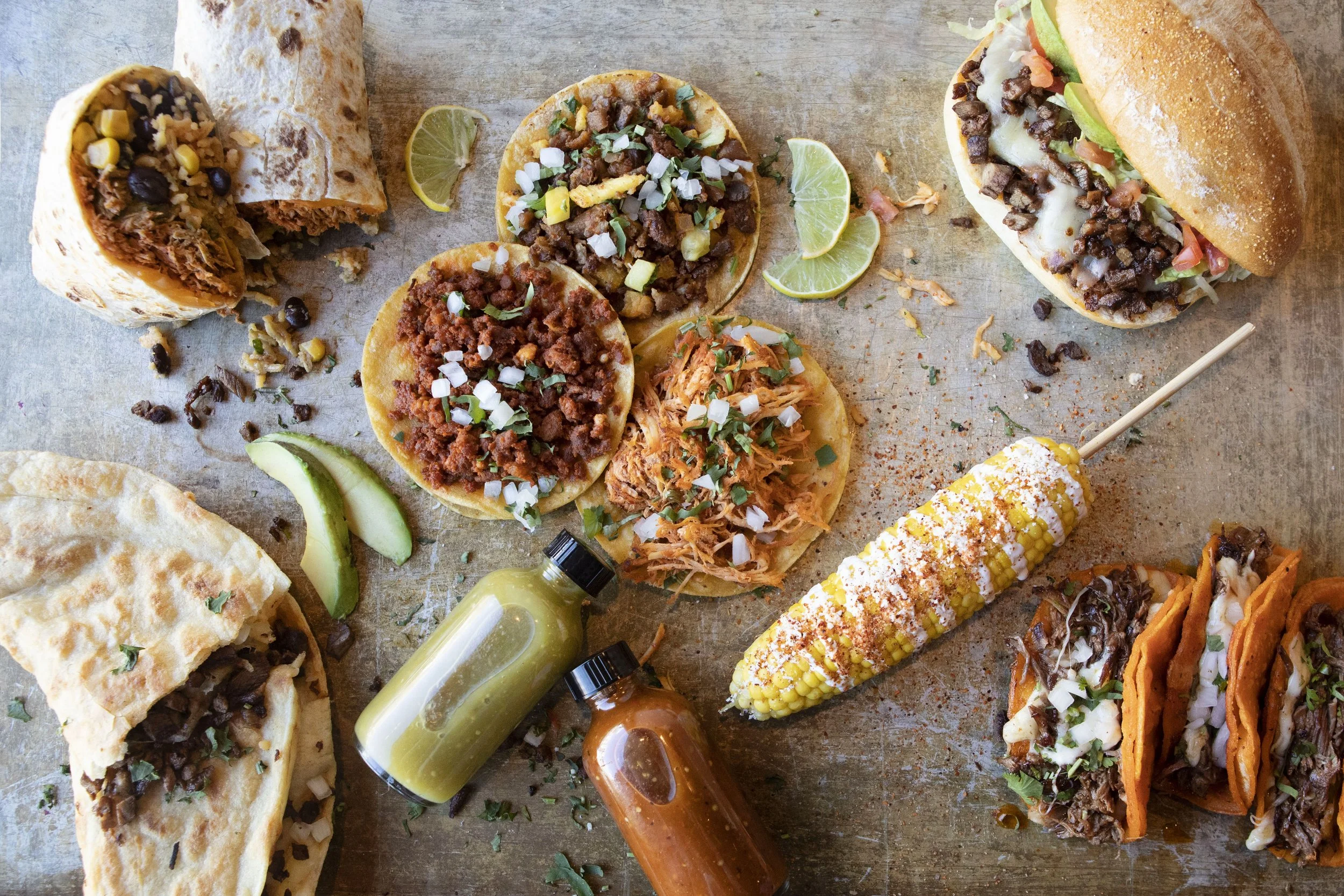 Assorted Mexican street tacos on a wooden surface with lime wedges and hot sauce bottles.