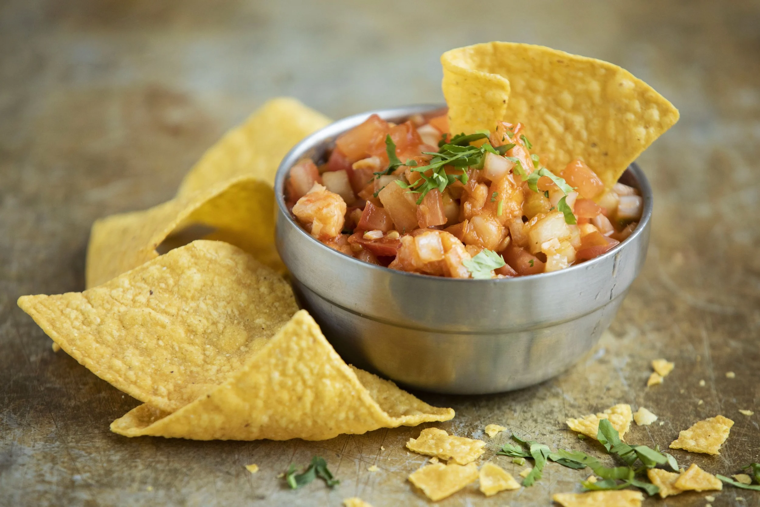 A metal bowl filled with shrimp ceviche garnished with cilantro, with yellow tortilla chips around it on a rustic surface.