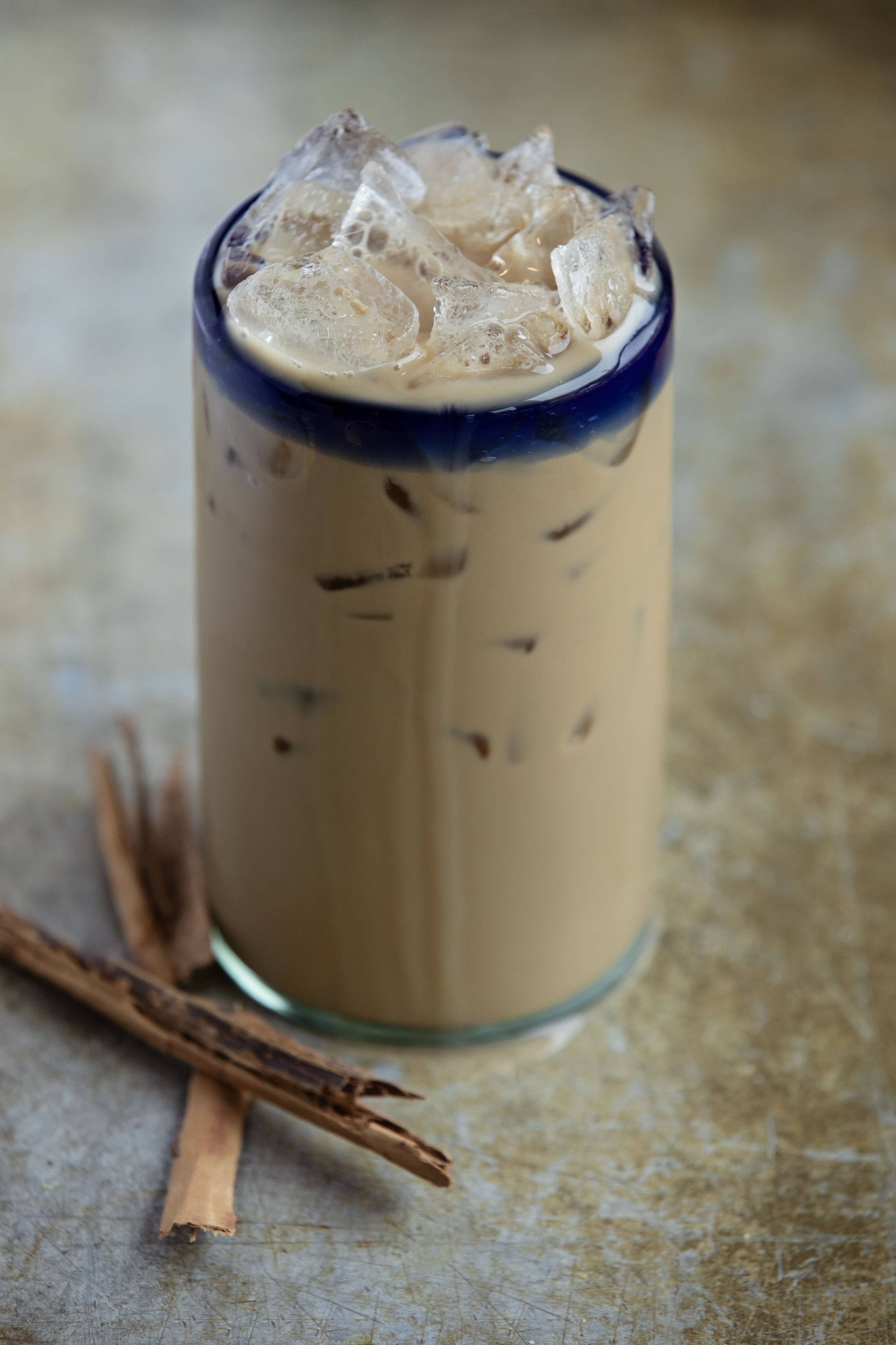 A glass of iced coffee topped with ice cubes, sitting on a textured surface with cinnamon sticks nearby.