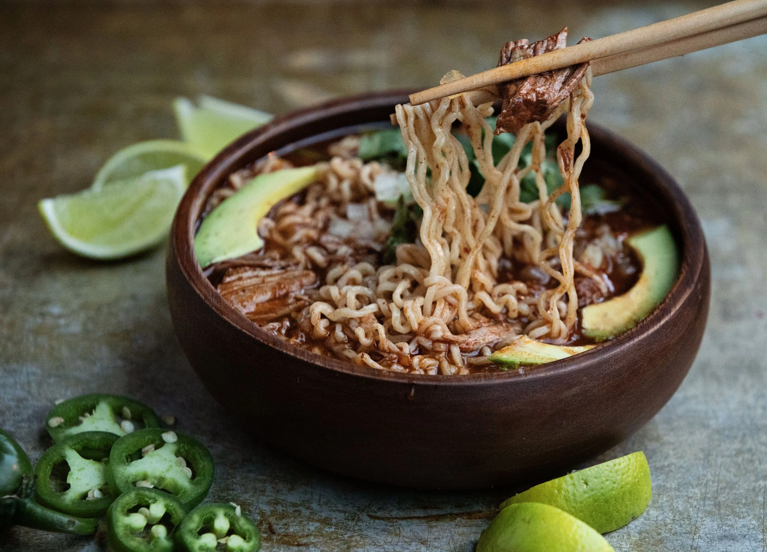 Close-up of a bowl of ramen noodles with slices of avocado and lime wedges, with lime slices and jalapeño peppers in the foreground.