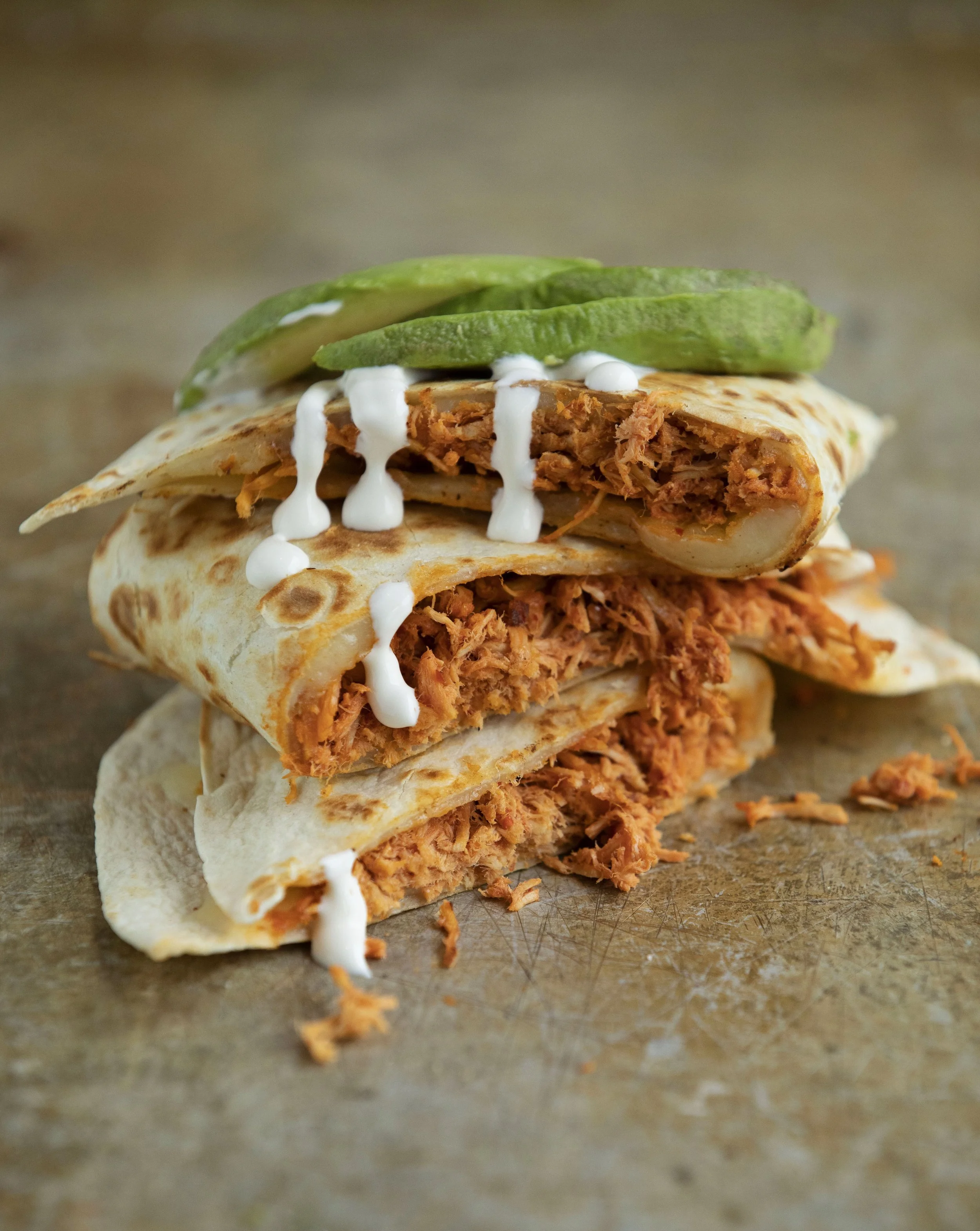 Close-up of a shredded chicken taco with white sauce and sliced avocado on top, resting on a brown surface.