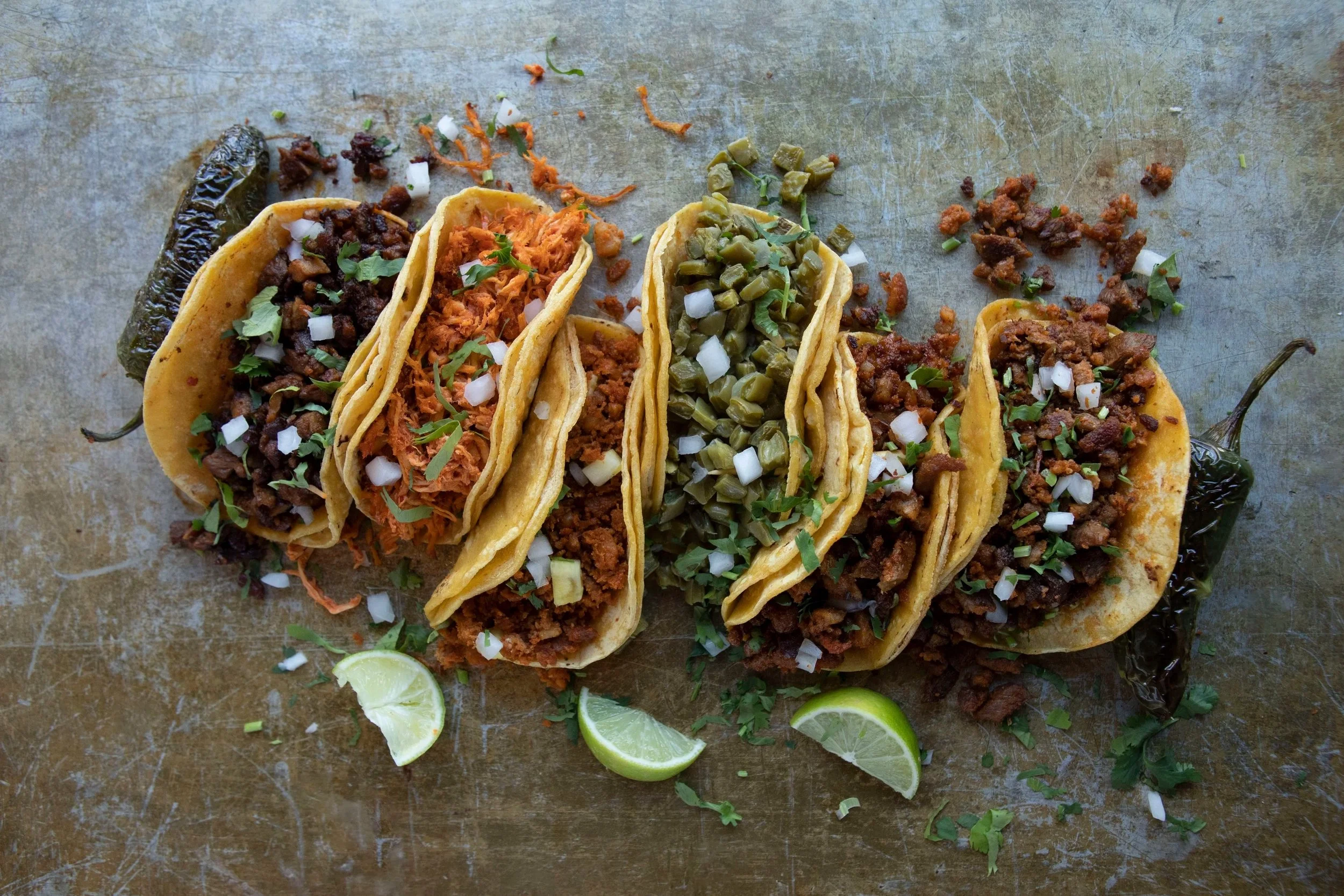 Five tacos filled with various meats, topped with chopped onions and fresh herbs, served on a rustic surface with lime wedges and scattered herbs around.