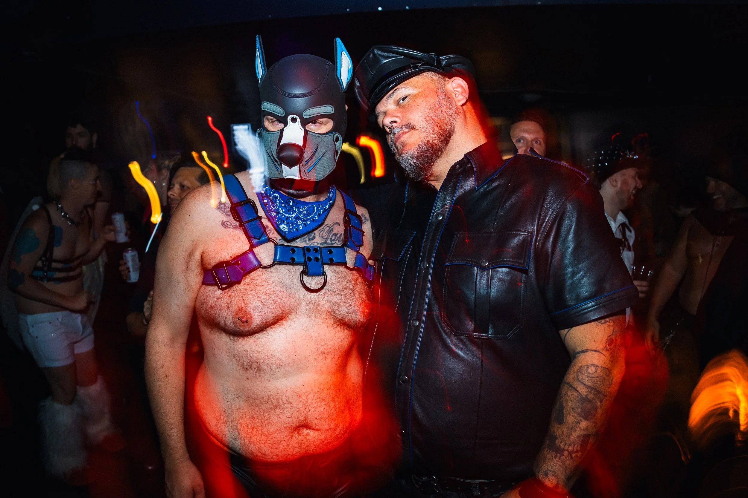 Two men at a party, one wearing a dog mask and blue bandana, shirtless with tattoos, and the other in a black leather jacket and goggles, surrounded by people in casual and themed costumes.