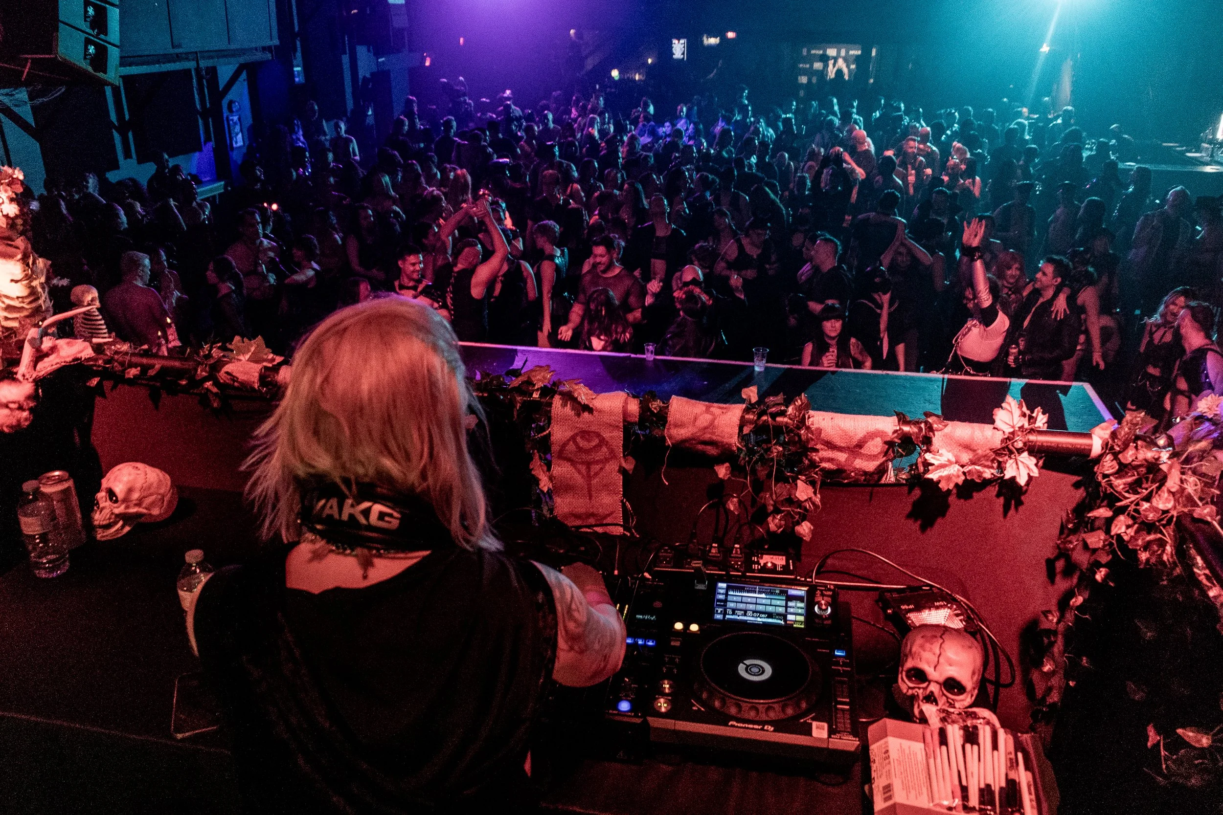 A large crowd of people dancing and socializing in a dark nightclub with purple and blue lighting. In the foreground, a DJ is operating a mixing console with skull decorations nearby.