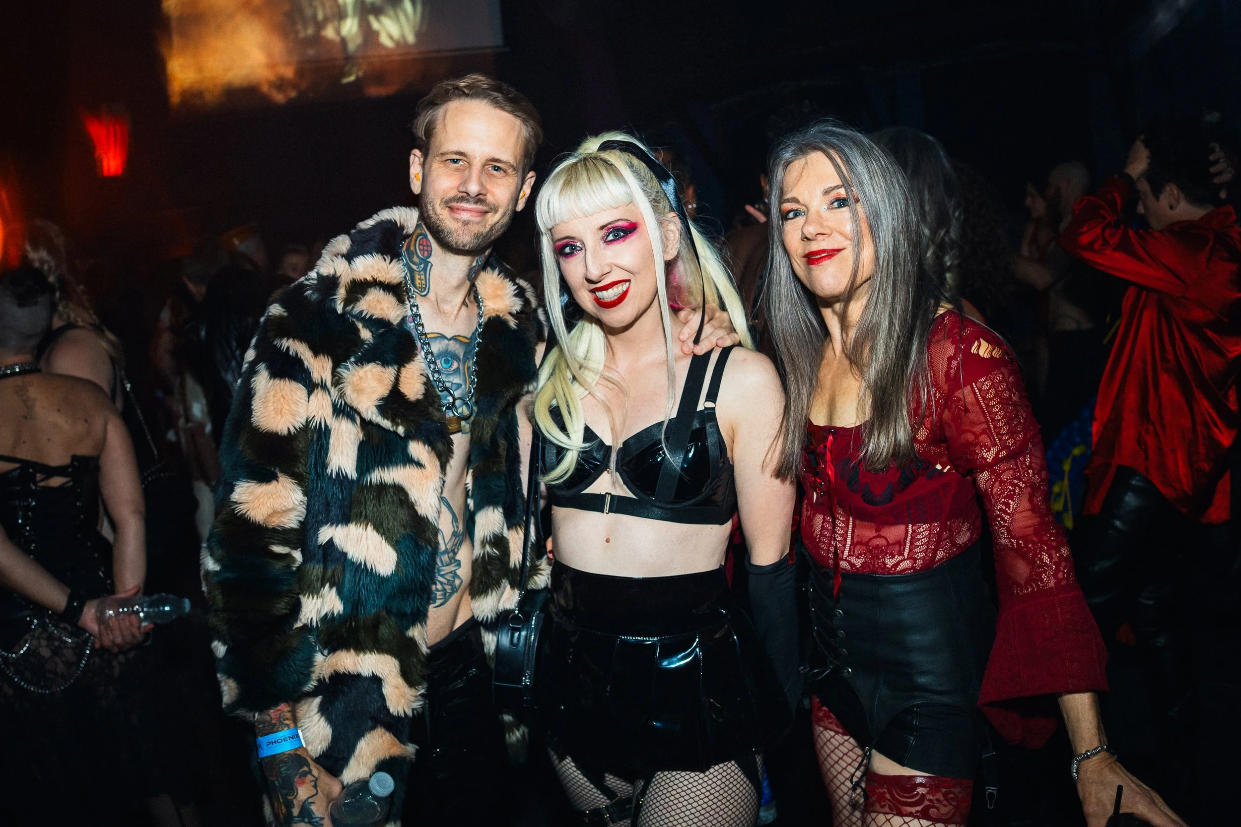 Three people dressed in alternative fashion at a party or nightclub. The man on the left has short hair, a beard, tattoos, and is wearing a fur coat with a chain necklace. The woman in the middle has platinum blonde hair with bangs, bold makeup, and 