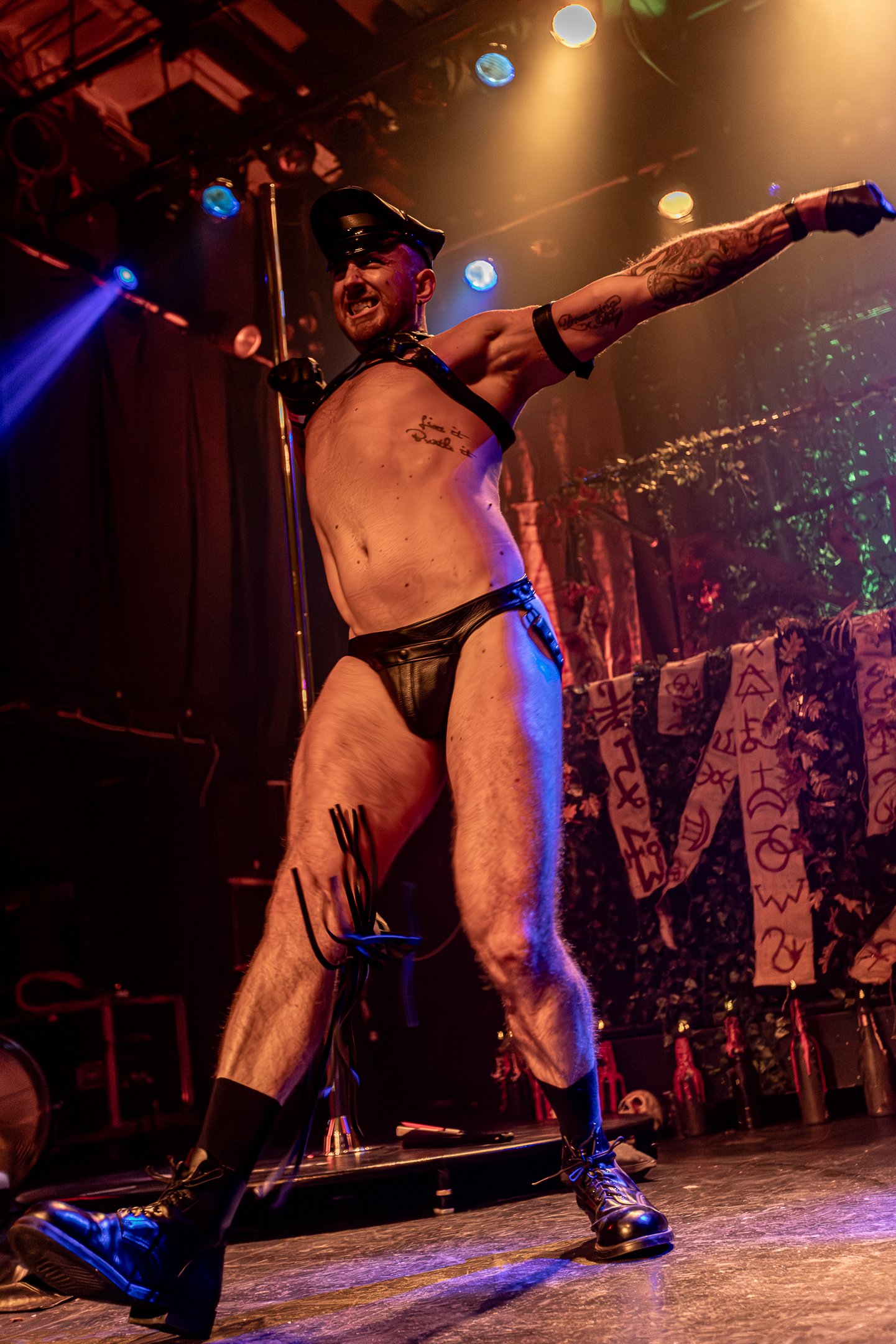 A muscular man in leather shorts and a police cap is performing a dance on stage with a pole, with colorful stage lights illuminating the scene and curtains in the background.
