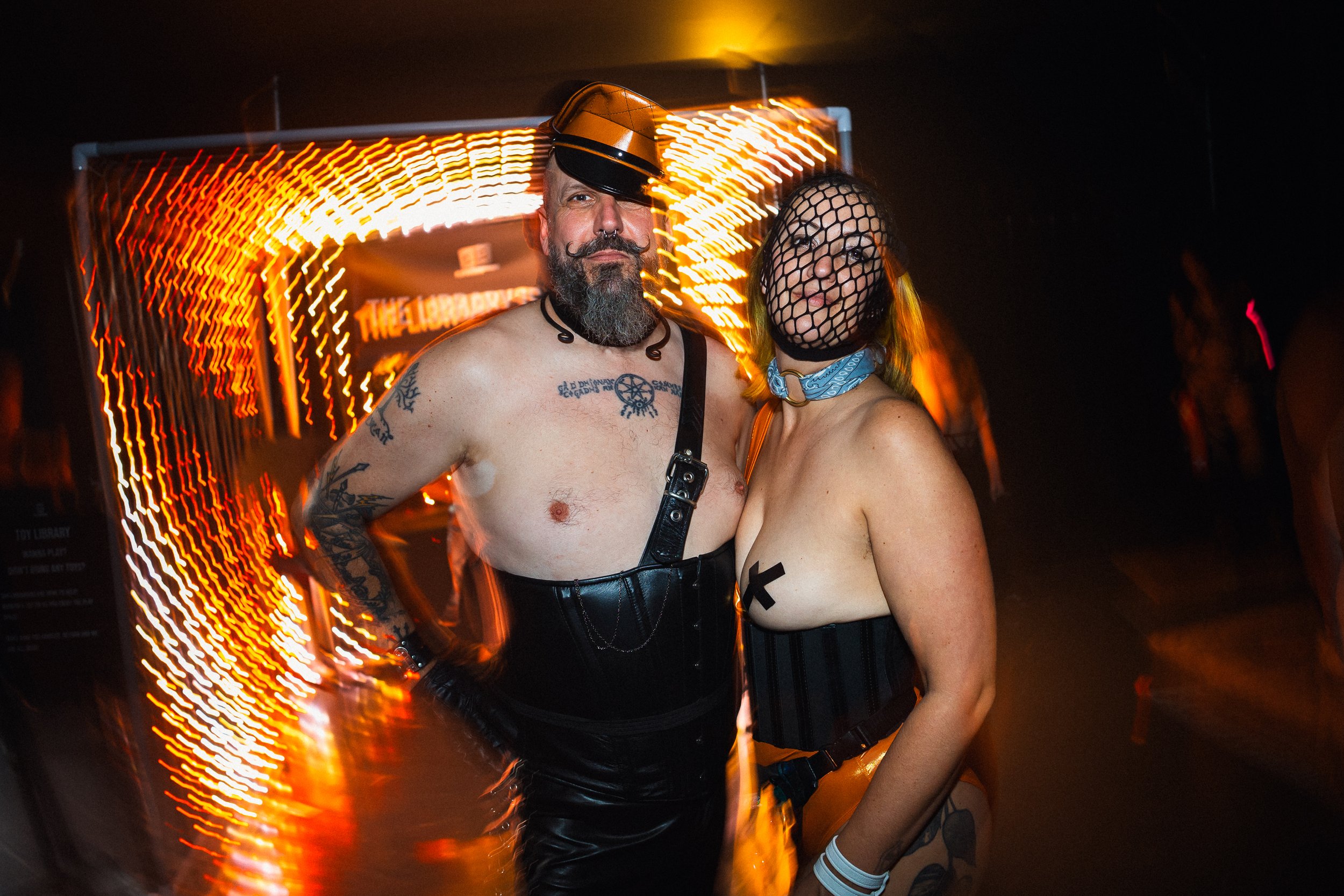 Two people dressed in BDSM or fetishwear posing together at night, with neon orange and yellow lights creating light trails in the background.
