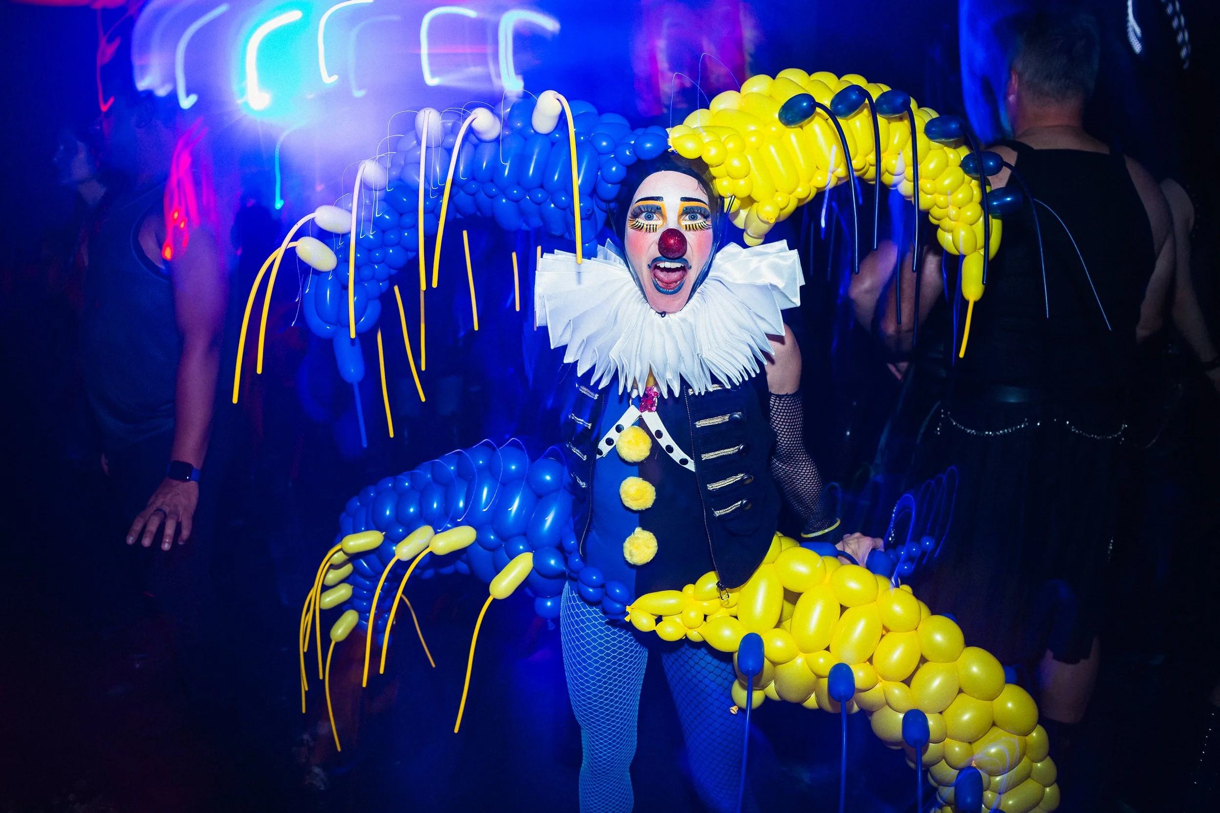 A person dressed as a colorful clown with a balloon costume, wearing a white ruffled collar, clown makeup, and a red nose, at a party or event with blue and neon lighting.