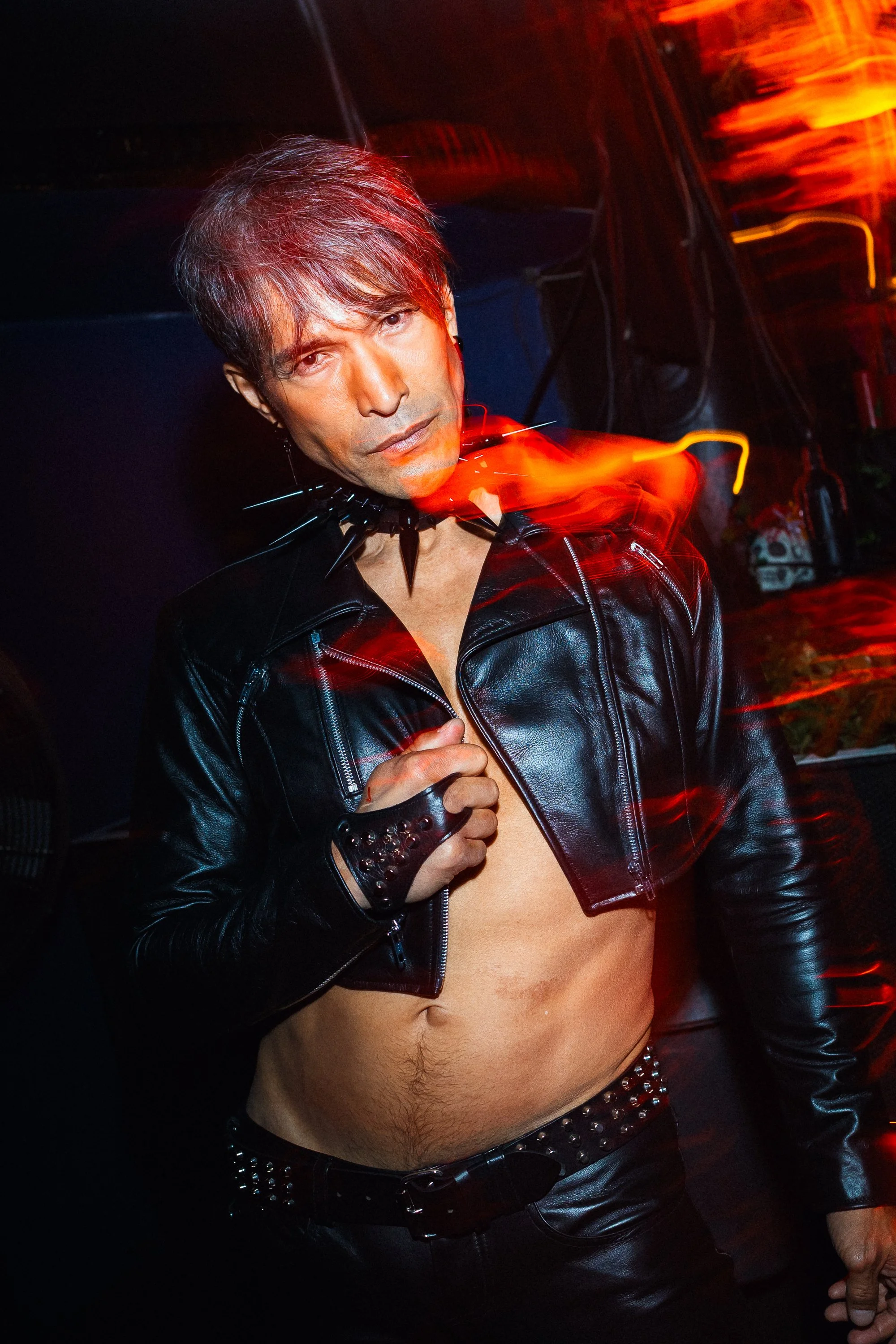 A person with short dark hair styled with a fringe, wearing a black leather jacket with silver spikes around the collar, a studded belt, and black leather pants, exposed midriff, standing in a dark environment with red and orange lighting, holding th