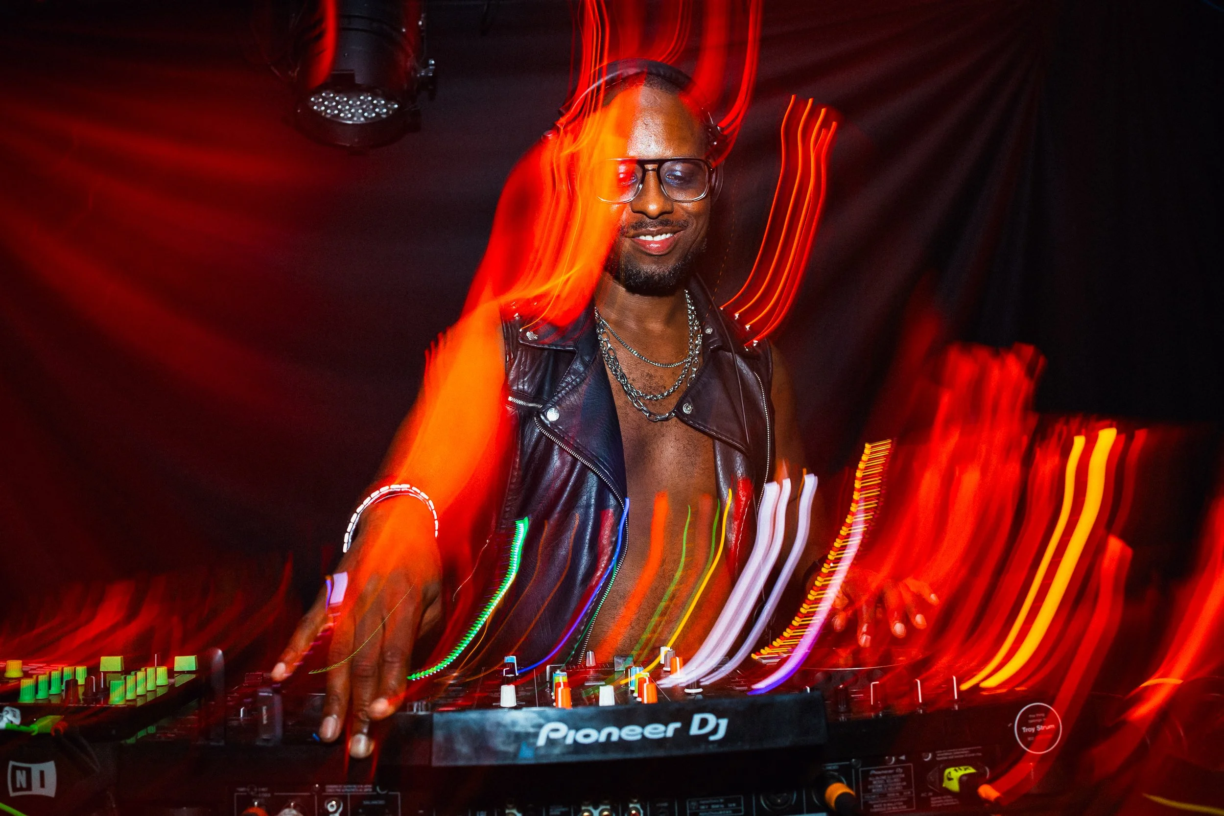 A DJ with glasses and a leather vest is mixing music on a Pioneer DJ controller with colorful lighting effects and motion blur.