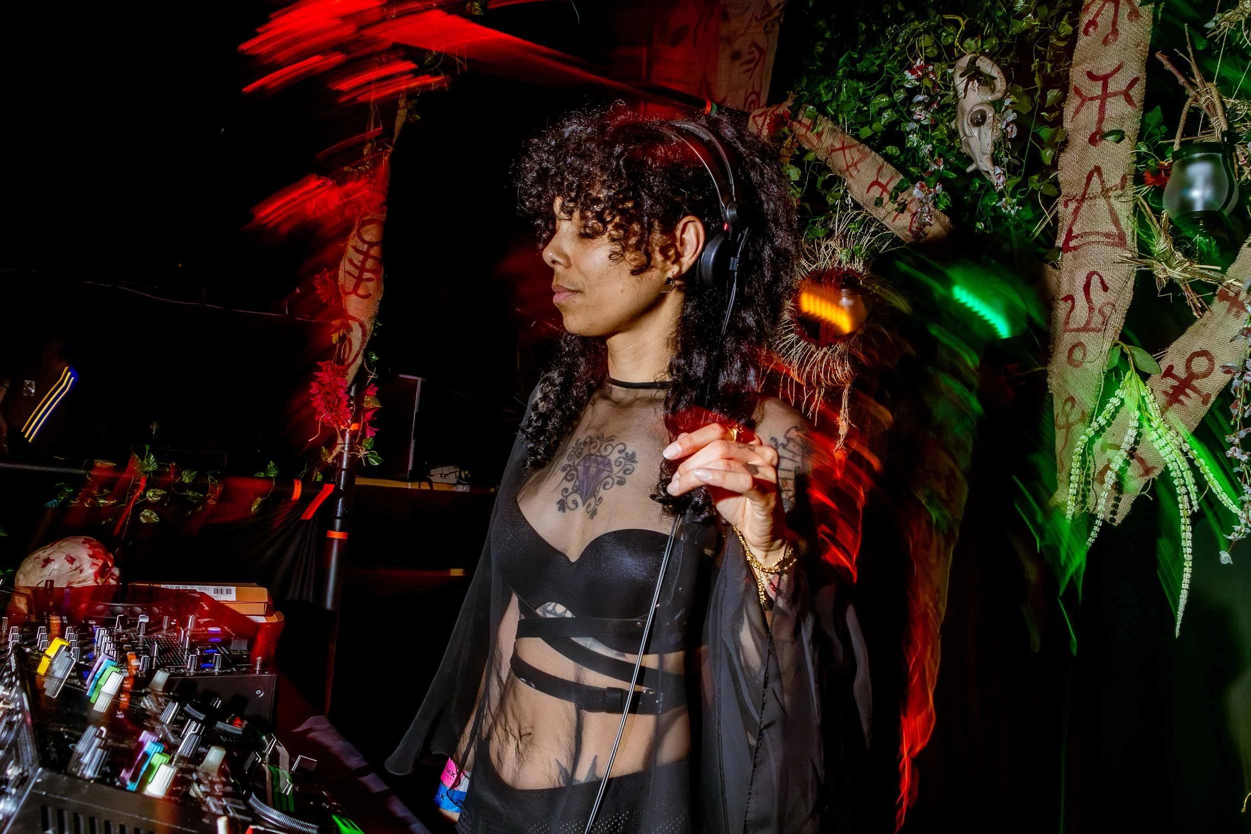 A female DJ with curly hair and tattoos, wearing headphones and a sheer black and striped outfit, is mixing music at a decorated party with greenery and red banners.