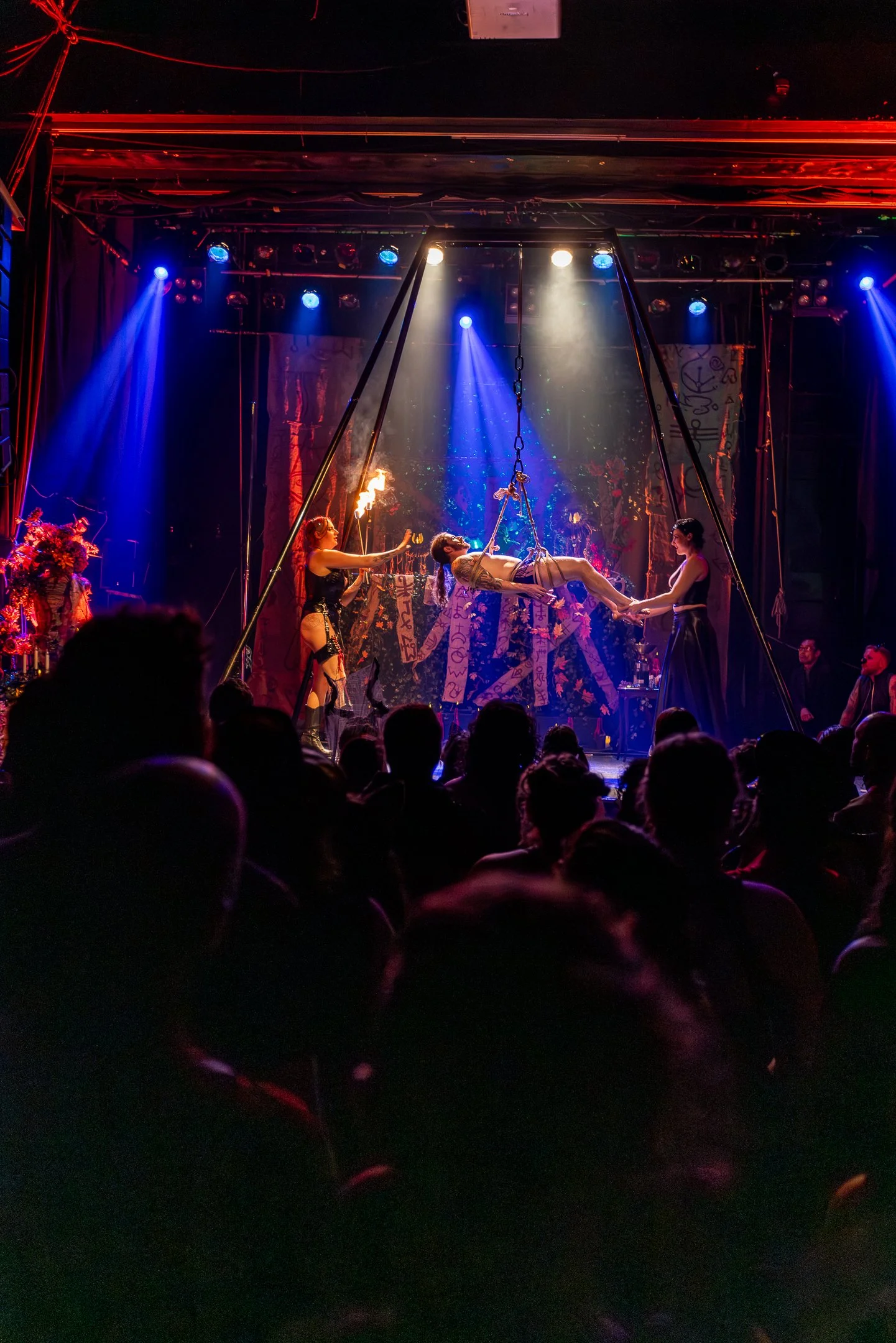 A circus or theater performance with performers on stage including a person holding a torch, another person on a swing, and another person in costume, under colorful stage lighting.