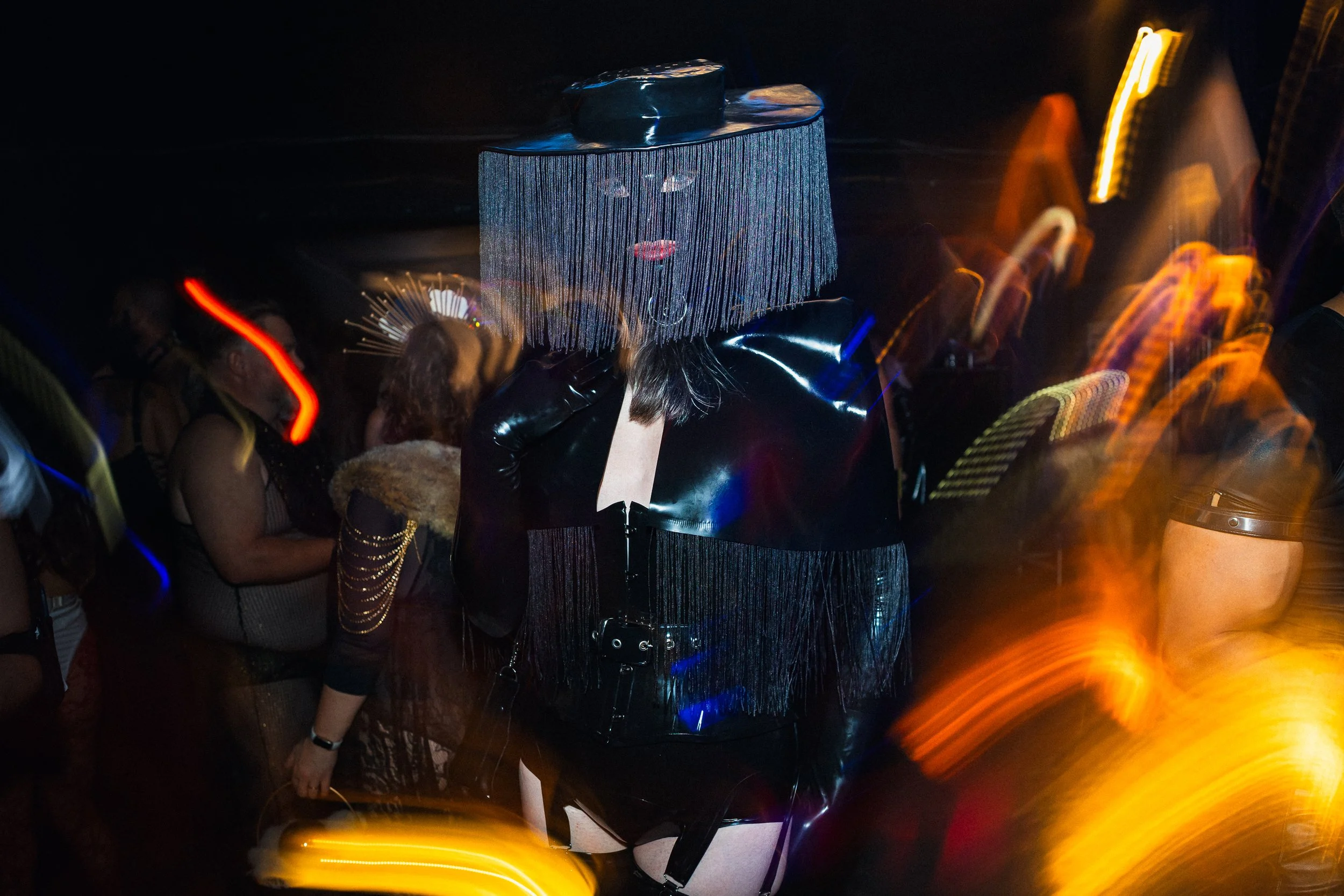 A person dressed in black with a striking headpiece made of fringes with a face print, lips, and eyes visible, standing among a crowd at a dark event with colorful light streaks.