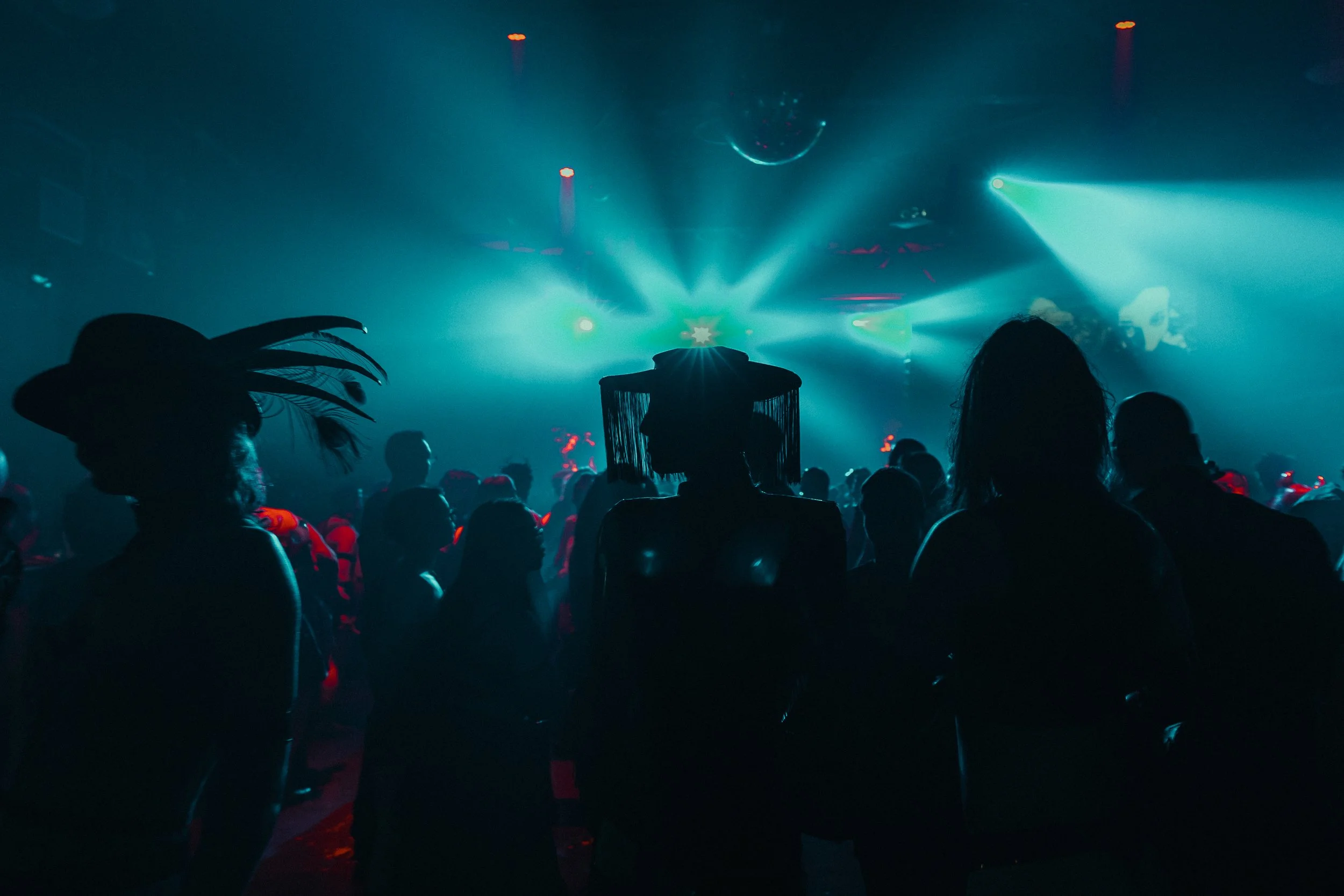 A crowded dance floor with silhouettes of people in a nightclub or concert venue, colorful lights, laser effects, and a disco ball overhead.
