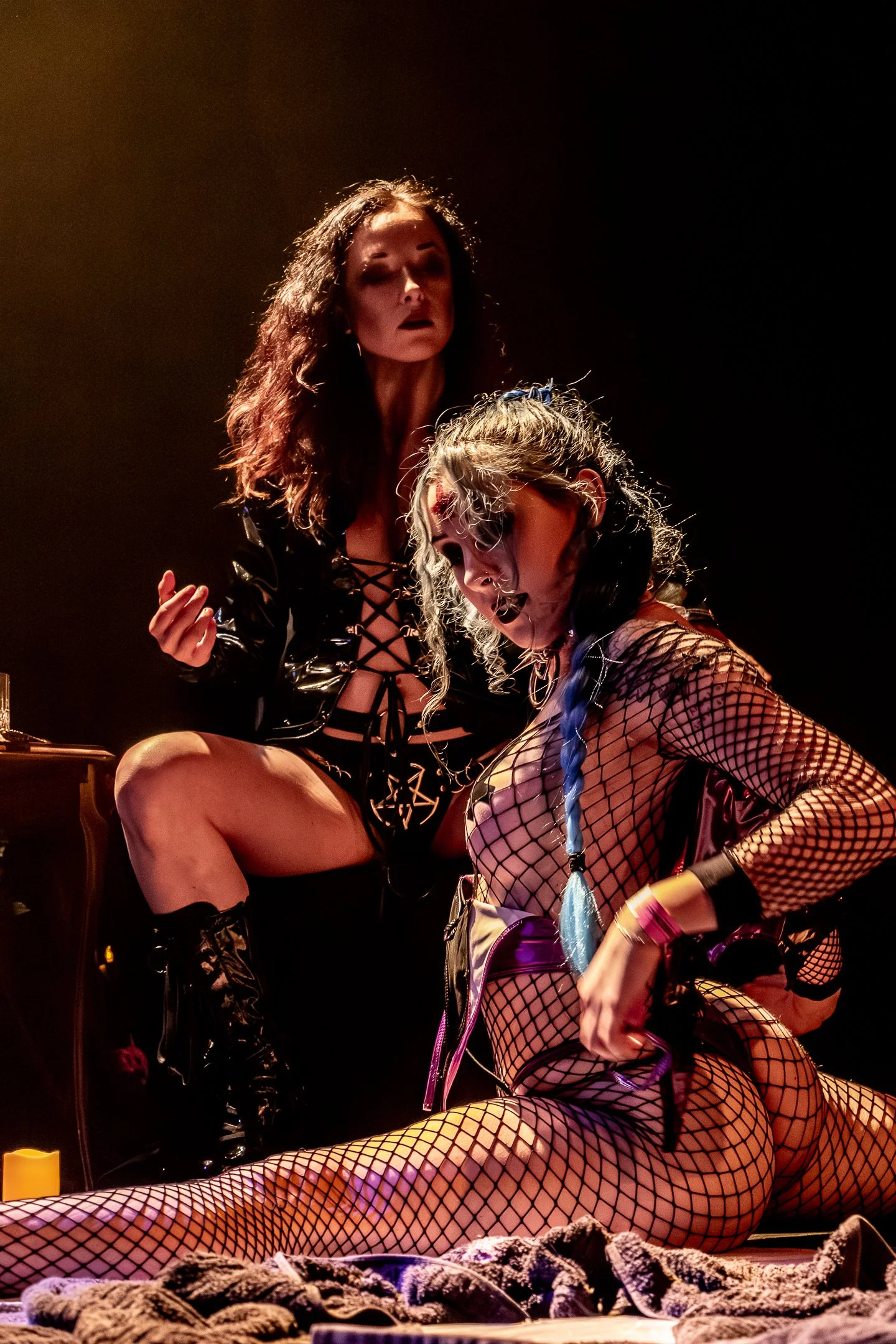 Two women with dramatic makeup and fishnet outfits are on stage in low lighting, with one woman sitting and the other standing over her, in a performance setting.