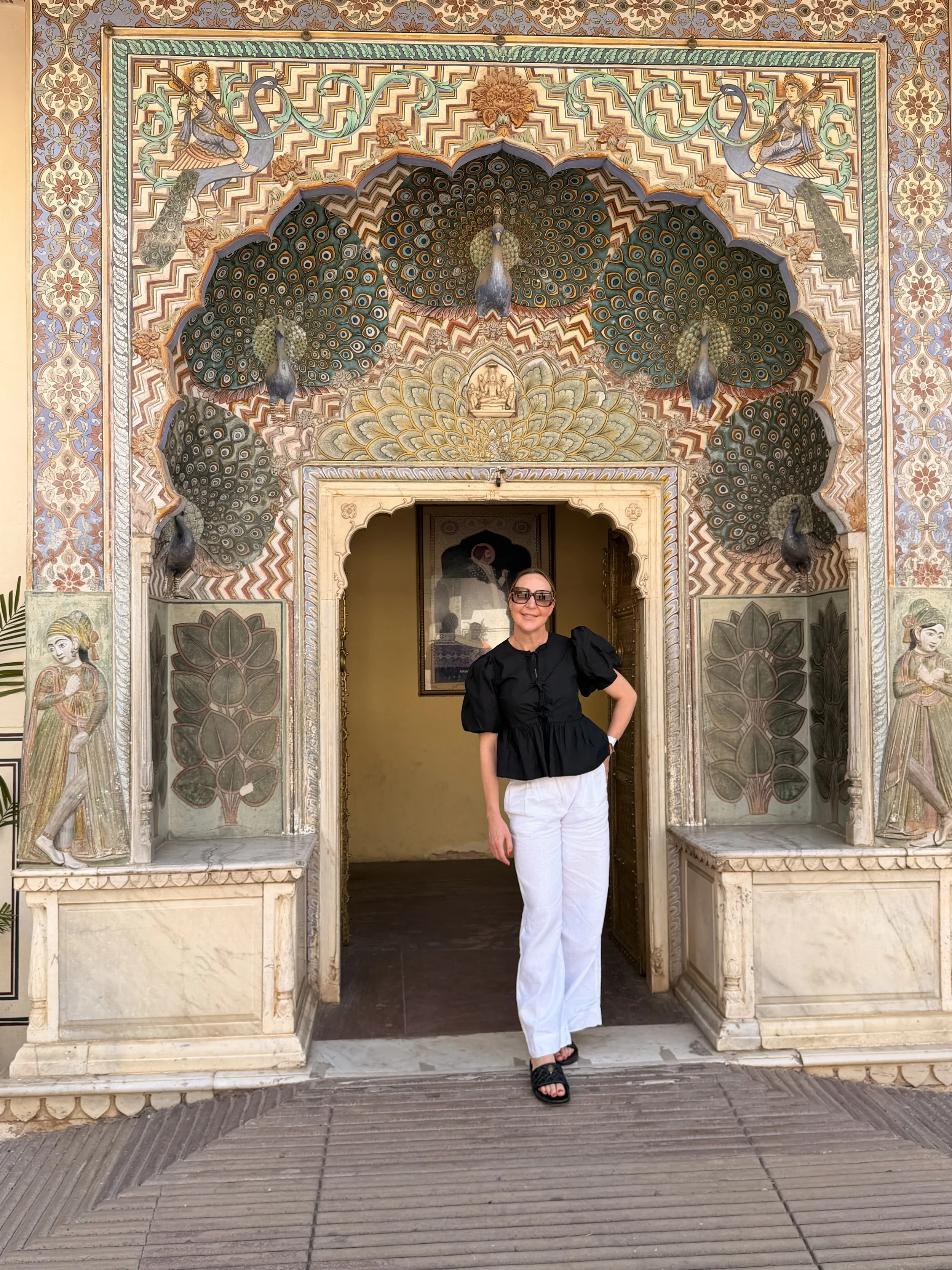 A woman with short hair, wearing sunglasses, a black top with puffed sleeves, and white pants, stands in front of an ornately decorated archway with peacock motifs and floral carvings. She is smiling with her hand on her hip.