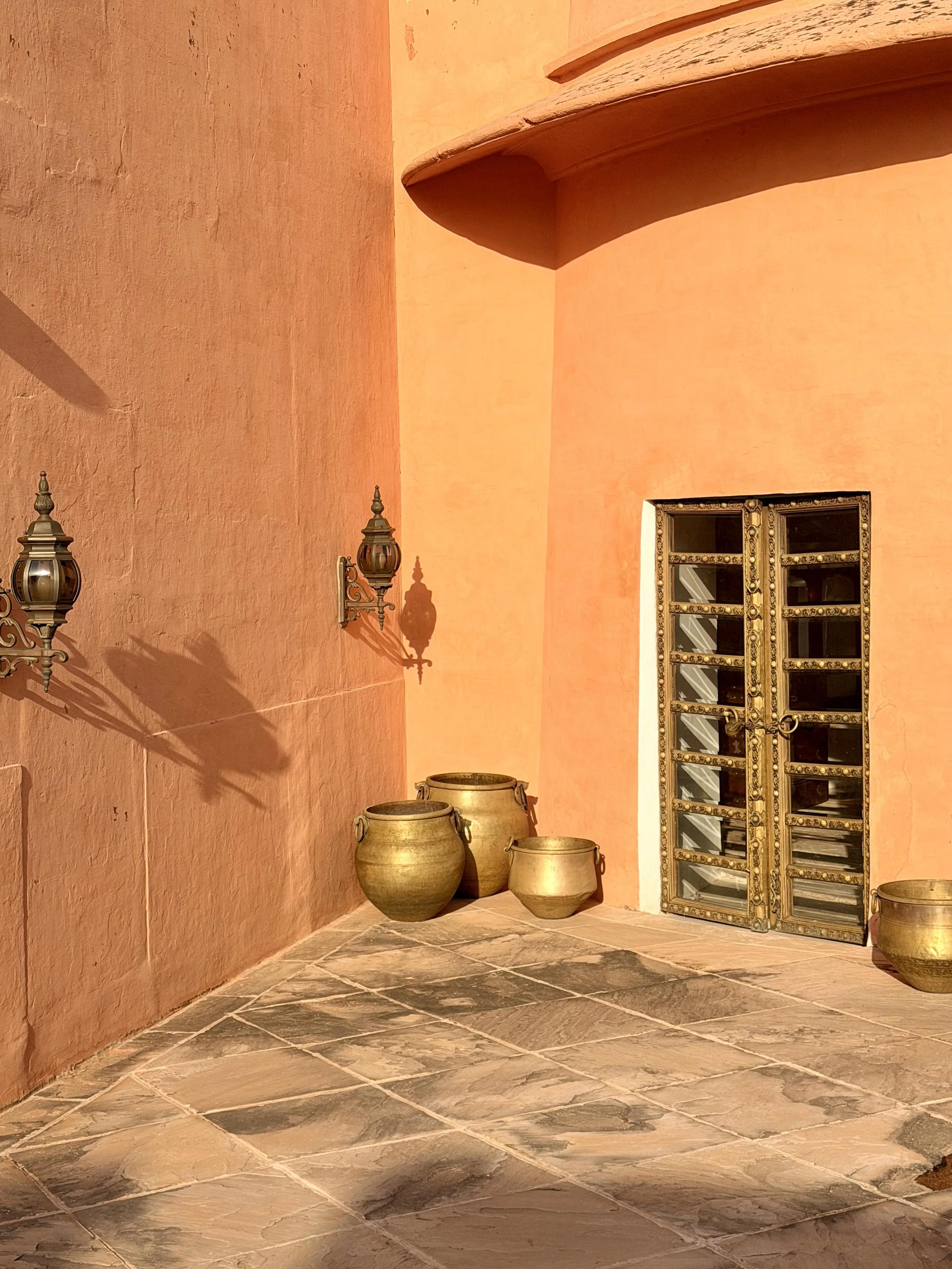 A corner of a building with pink and peach textured walls, two vintage-style lamps on the wall, three large ceramic pots on the ground, a window with ornate gold Trim, and a stone tile floor.