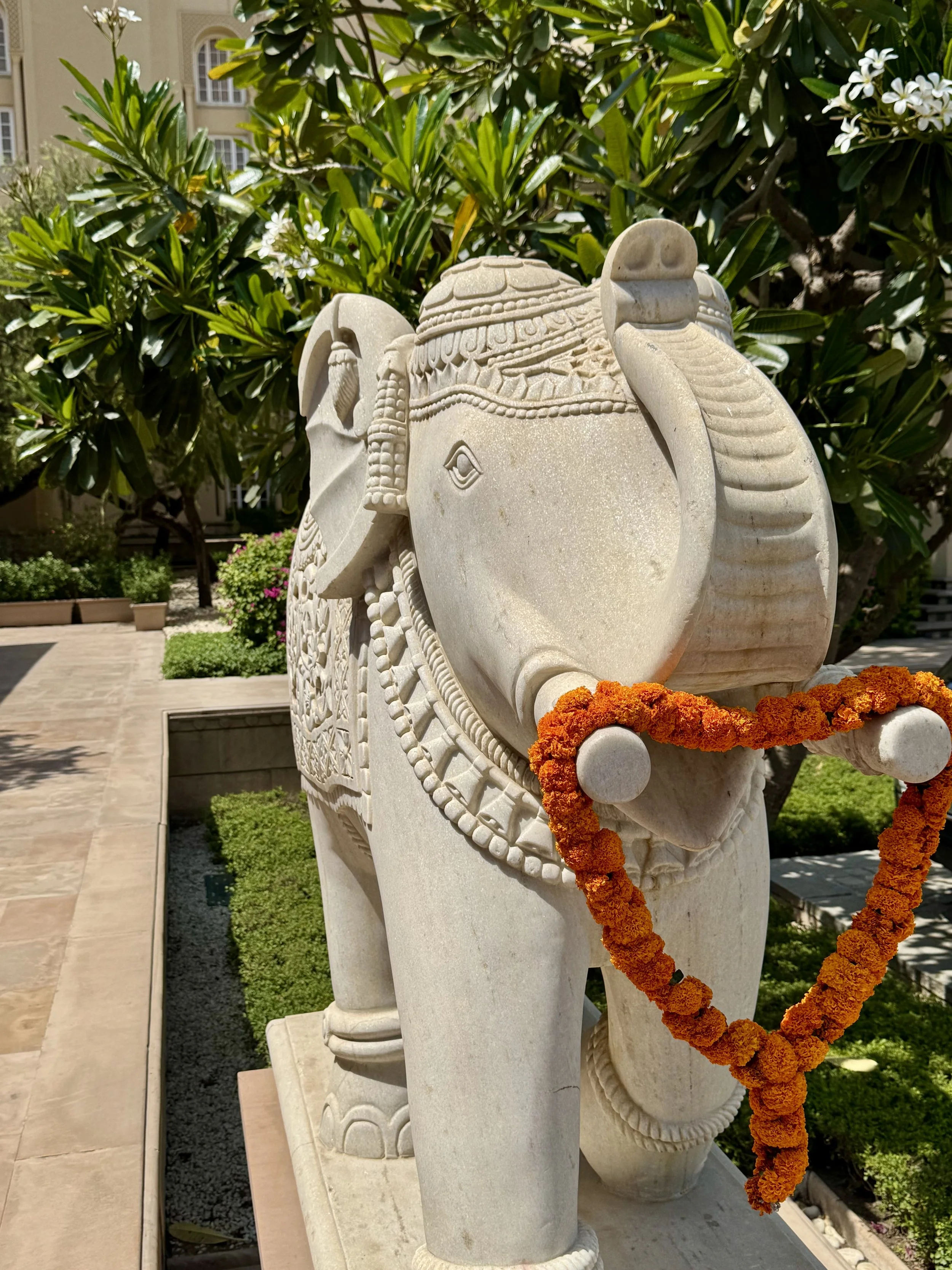 A stone elephant sculpture decorated with a marigold flower garland, set outdoors in a landscaped area with green bushes and a building in the background.