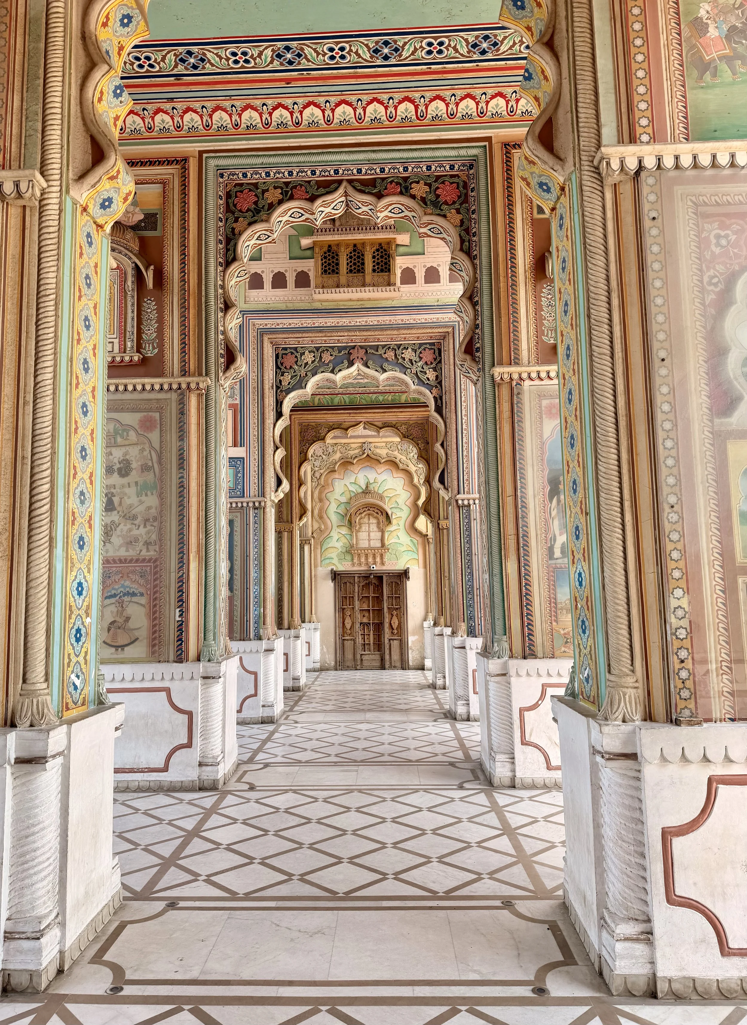 A corridor in a palace with intricately painted walls and ceilings, archways, and a tiled floor, leading to a wooden door at the end.