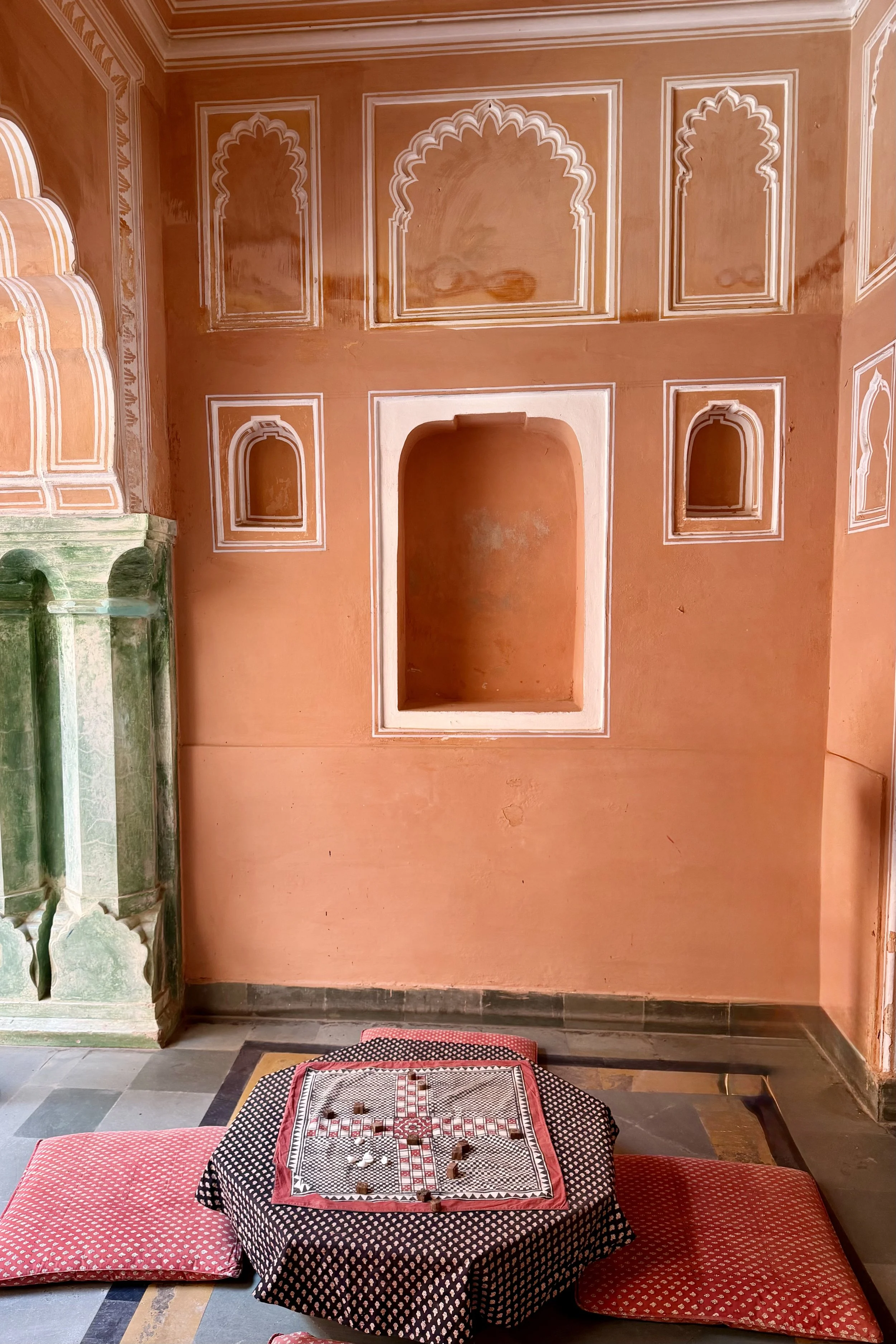 Interior of a room with orange walls decorated with white geometric patterns, an open archway, and small alcoves. A table with a chessboard and pieces is placed on a black and white patterned cloth, surrounded by red cushions, and a green marble colu