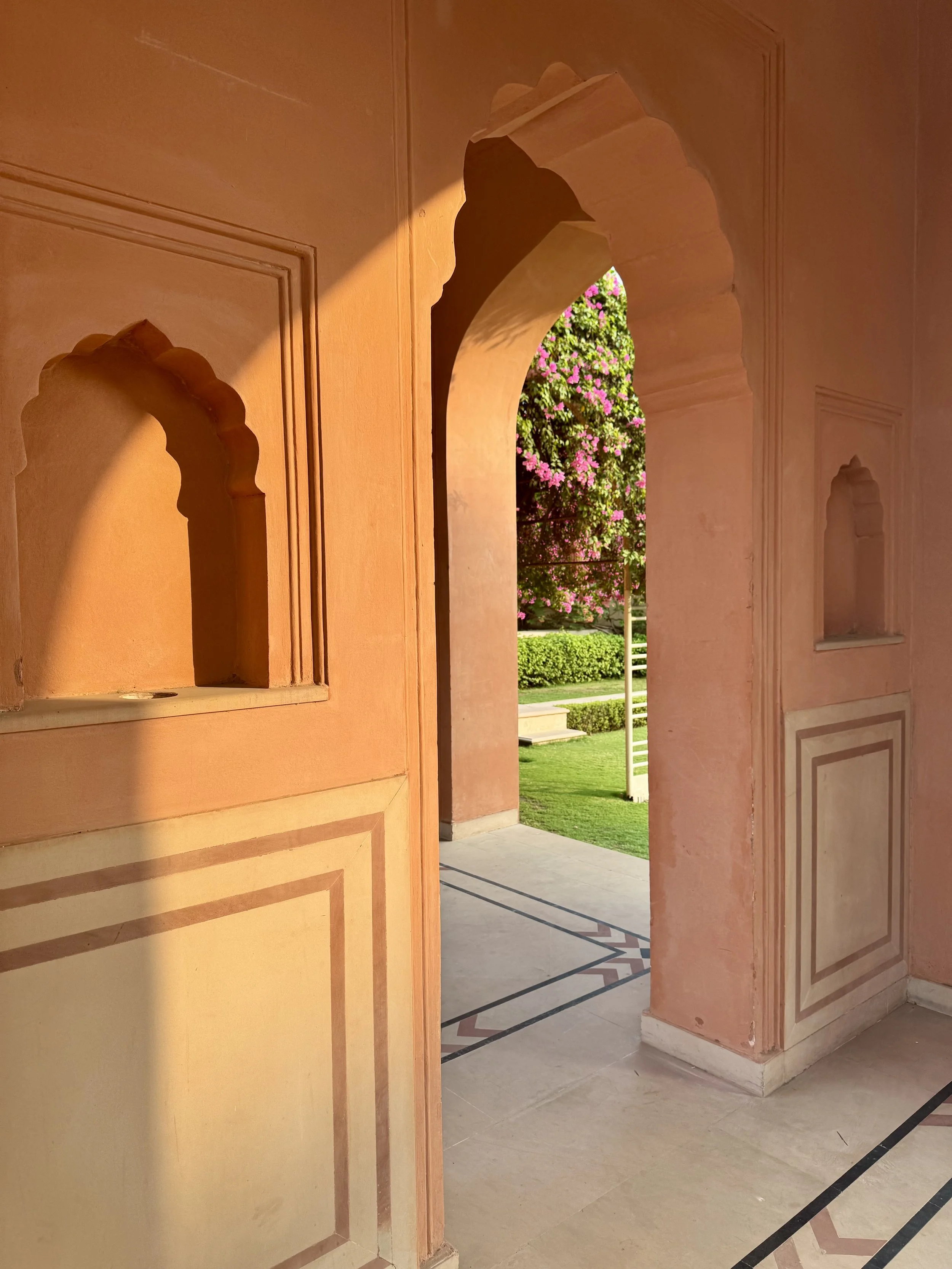 View of a corridor with peach-colored walls and decorative arches, leading to an outdoor garden with pink flowers and green bushes.