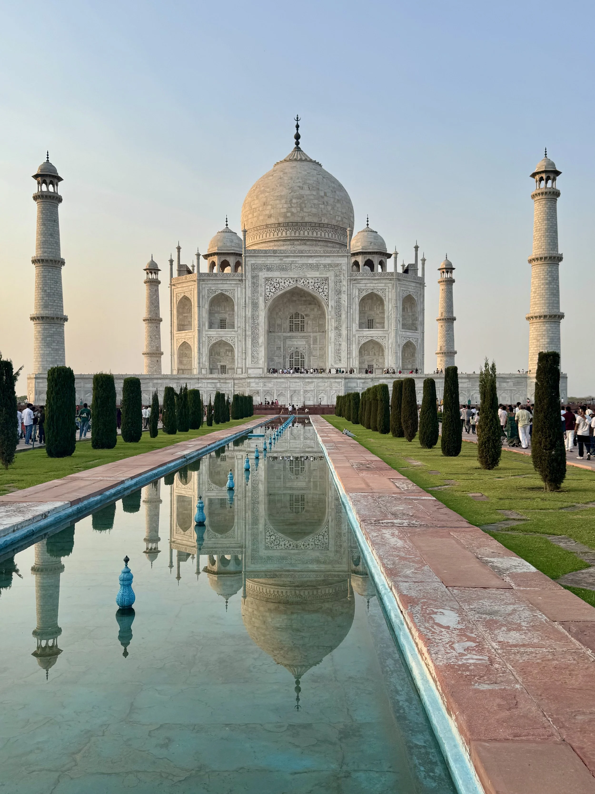 The Taj Mahal, a white marble mausoleum in India, reflecting in a long, narrow pool with a garden in the foreground and visitors walking around.