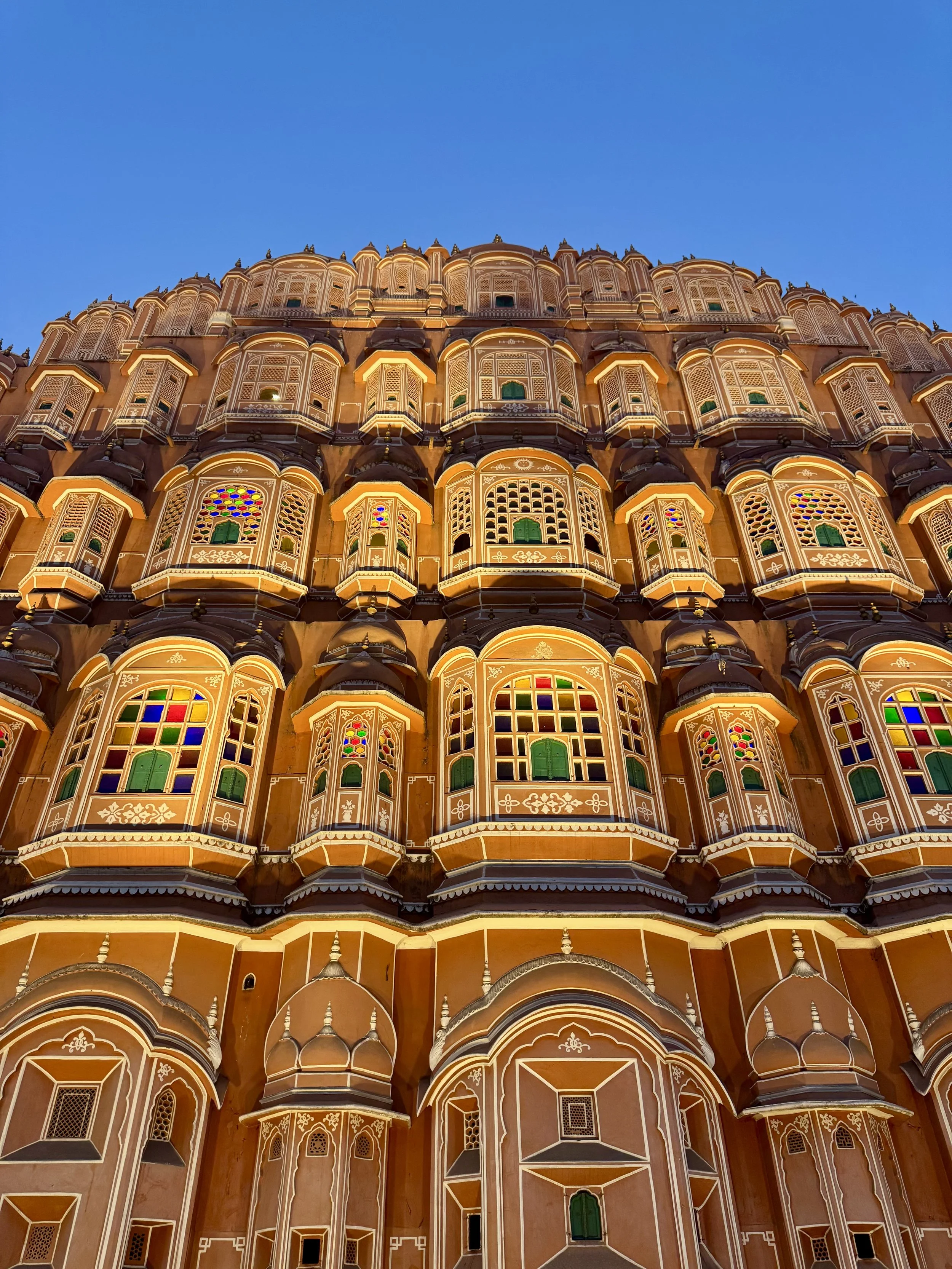 The image shows the front view of the Palace of the Winds in Jaipur, India, with detailed lattice windows and intricate architectural design illuminated in sunlight against a clear blue sky.
