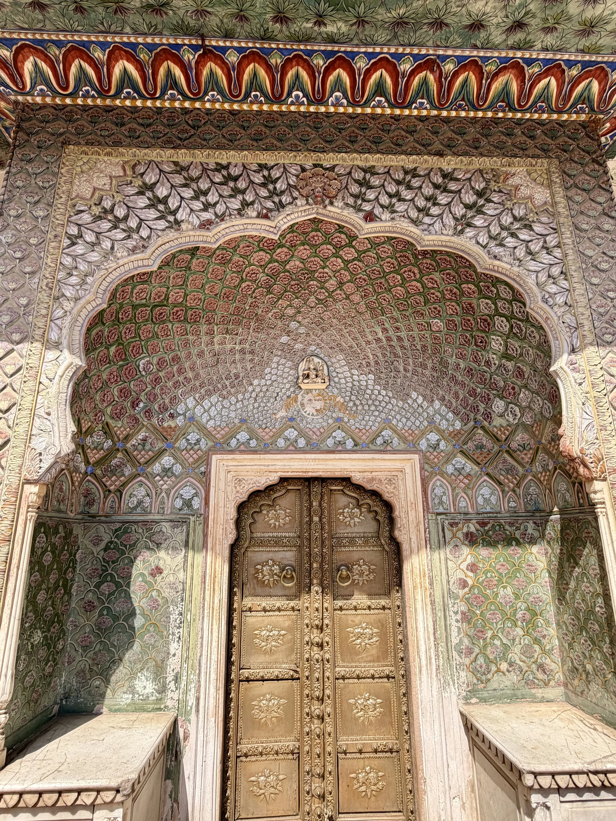 Decorative wooden door with golden carvings and floral motifs, surrounded by intricate painted arches and a beautifully painted ceiling with floral patterns in pink, green, and gold hues.