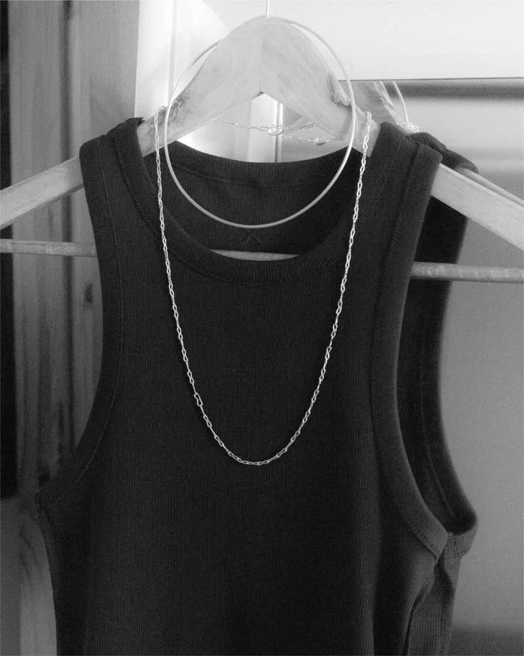 A black sleeveless top hanging on a wooden hanger with a necklace draped over it in a grayscale photo.