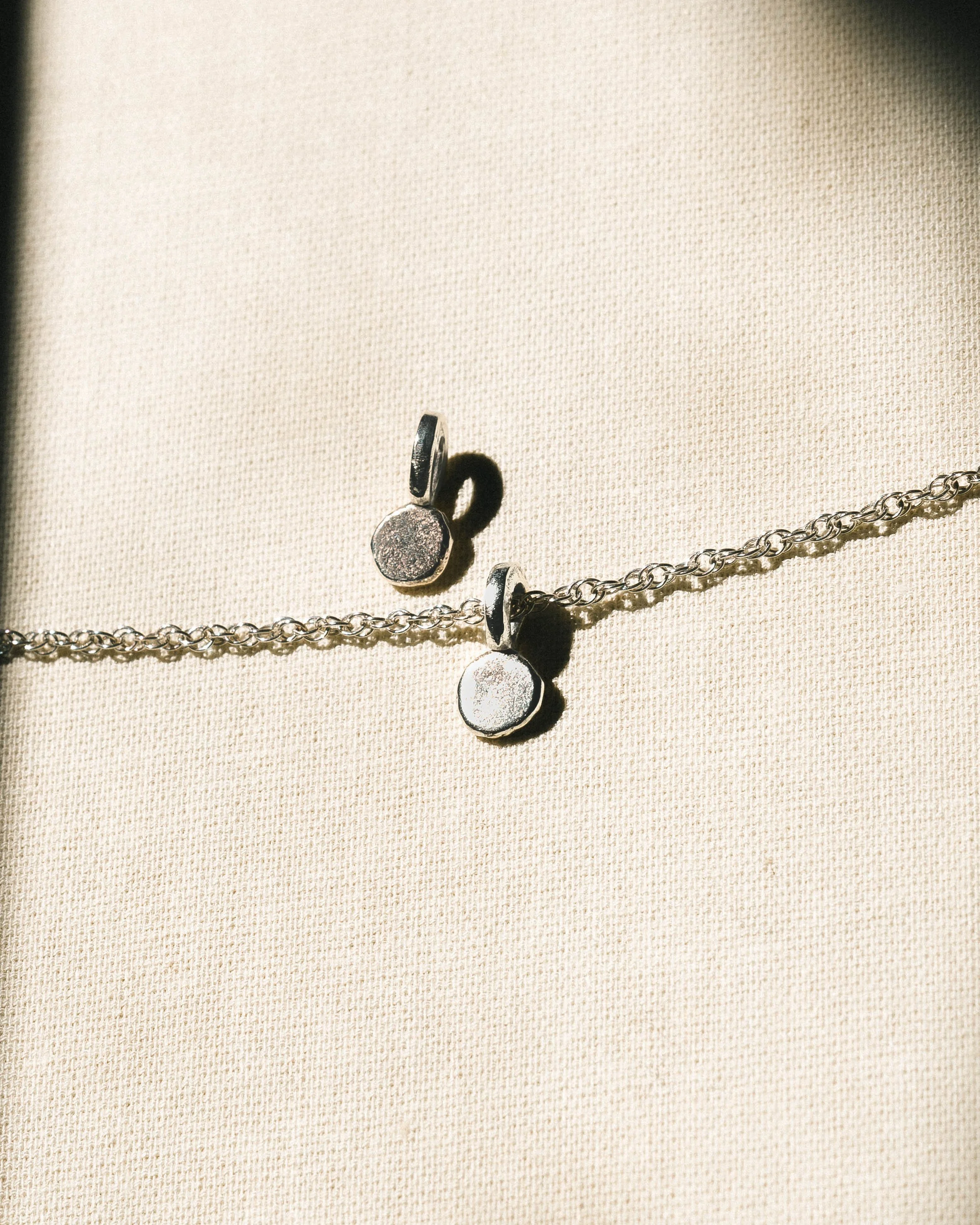 Close-up of silver jewelry pieces, including a chain and small round charms, laid on a light fabric background.