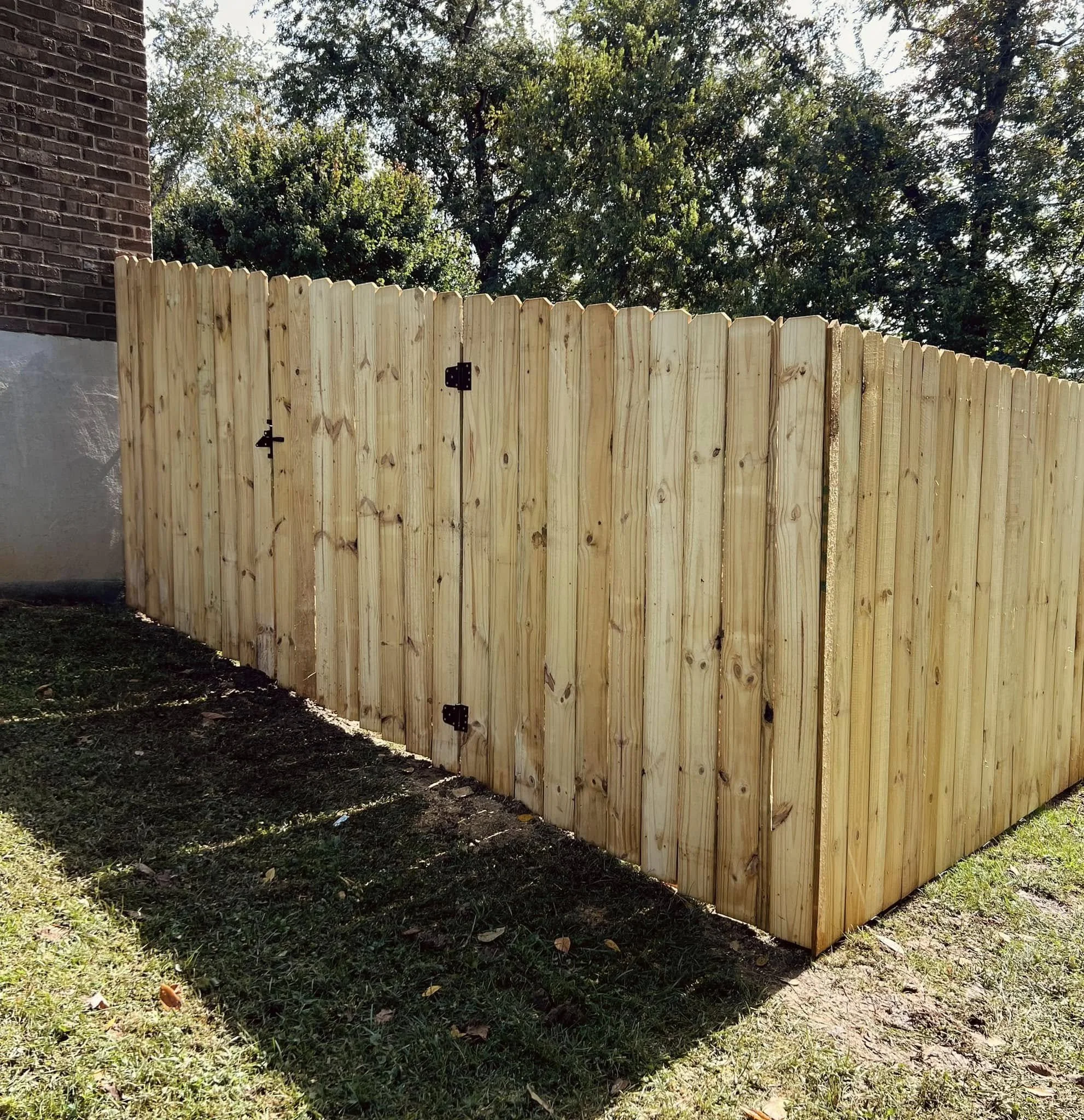 New wooden privacy fence with a latch, in front of trees and part of a brick house wall.